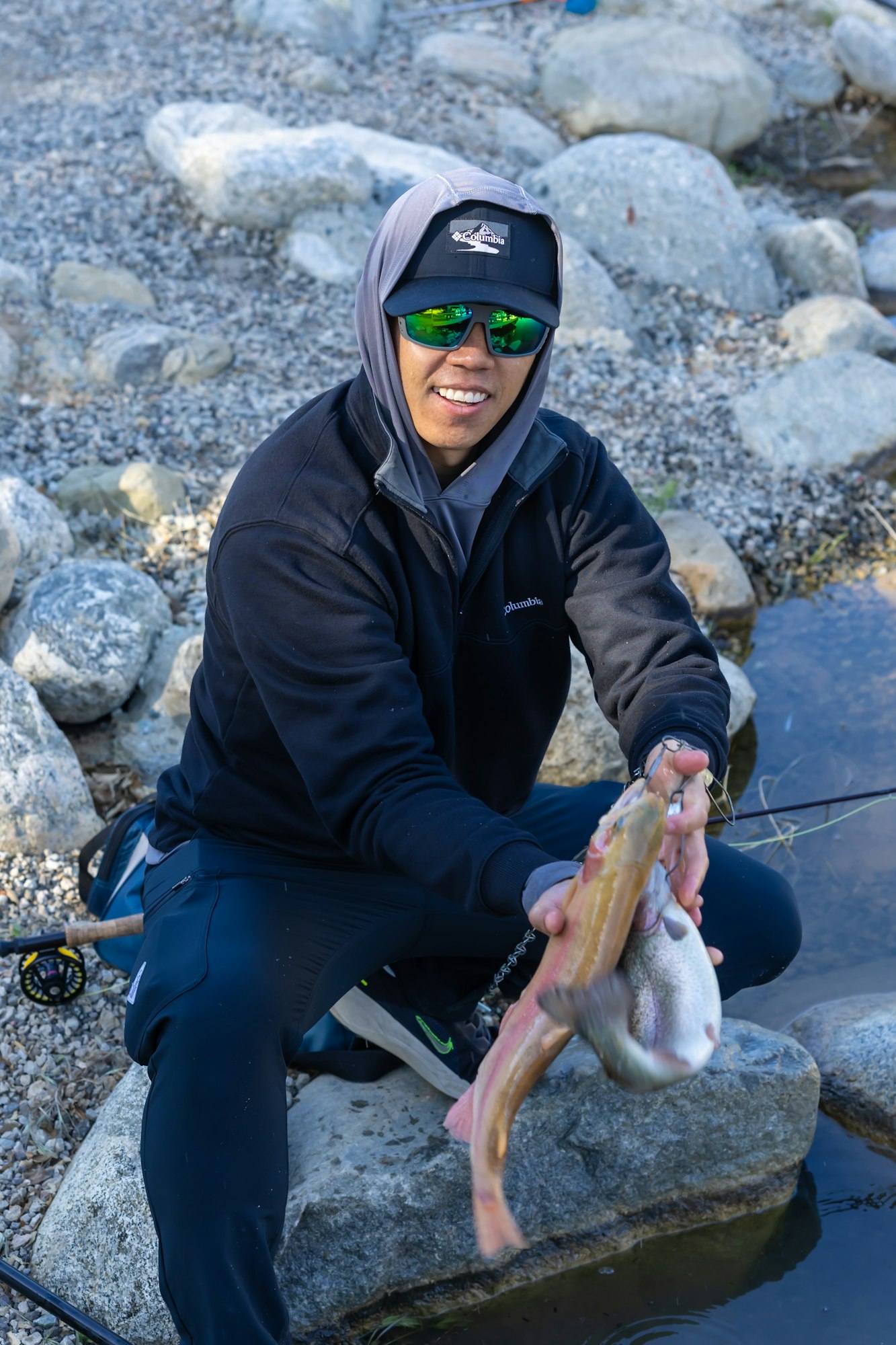 Person in a hoodie and sunglasses holding a fish by a rocky stream.