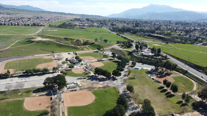 An aerial view of a green park with baseball fields, roadways, and residential areas, set against a mountainous backdrop.