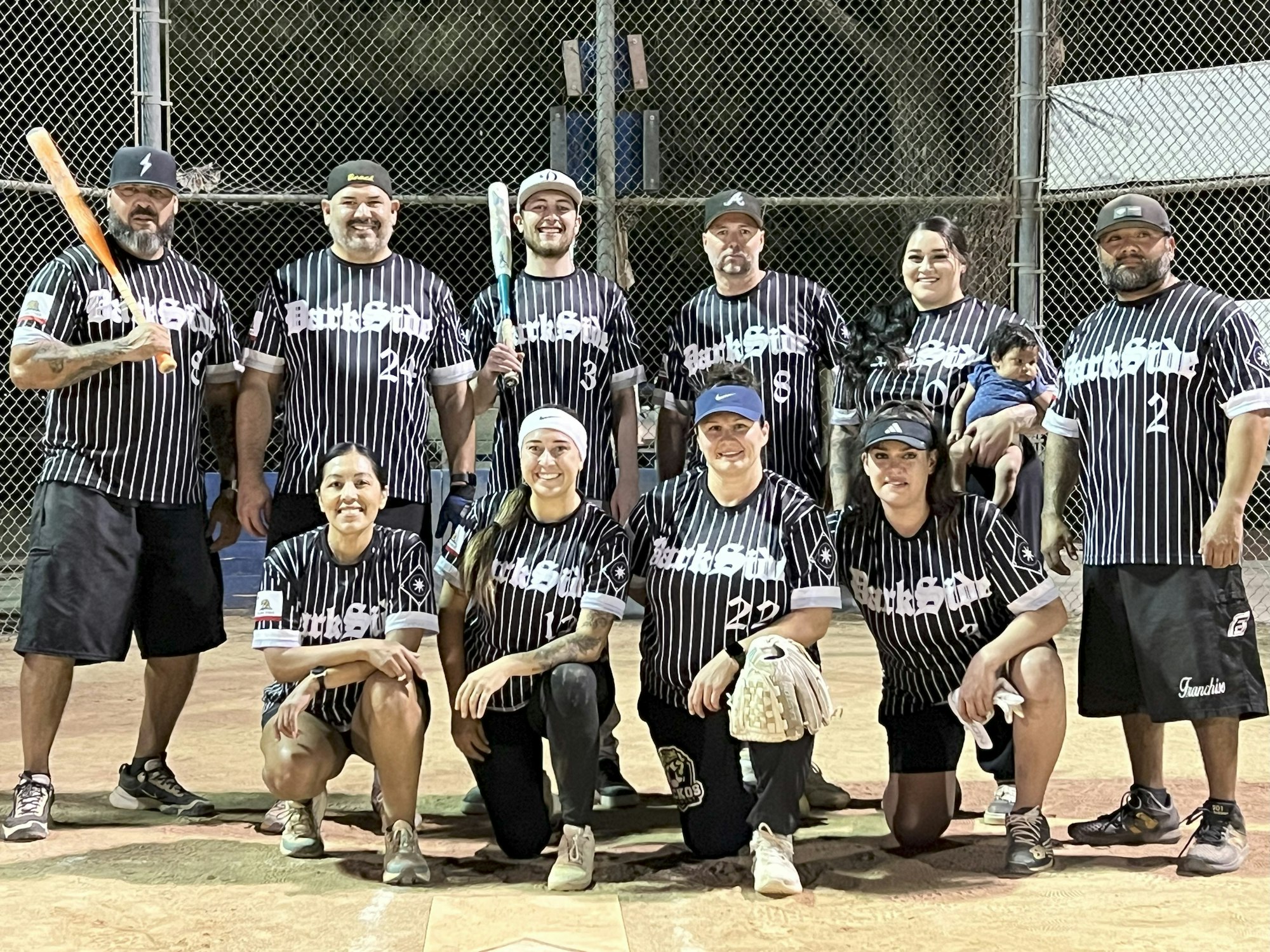 A softball team poses on the field. Players wear matching black and white striped jerseys and shorts, holding bats and gloves.