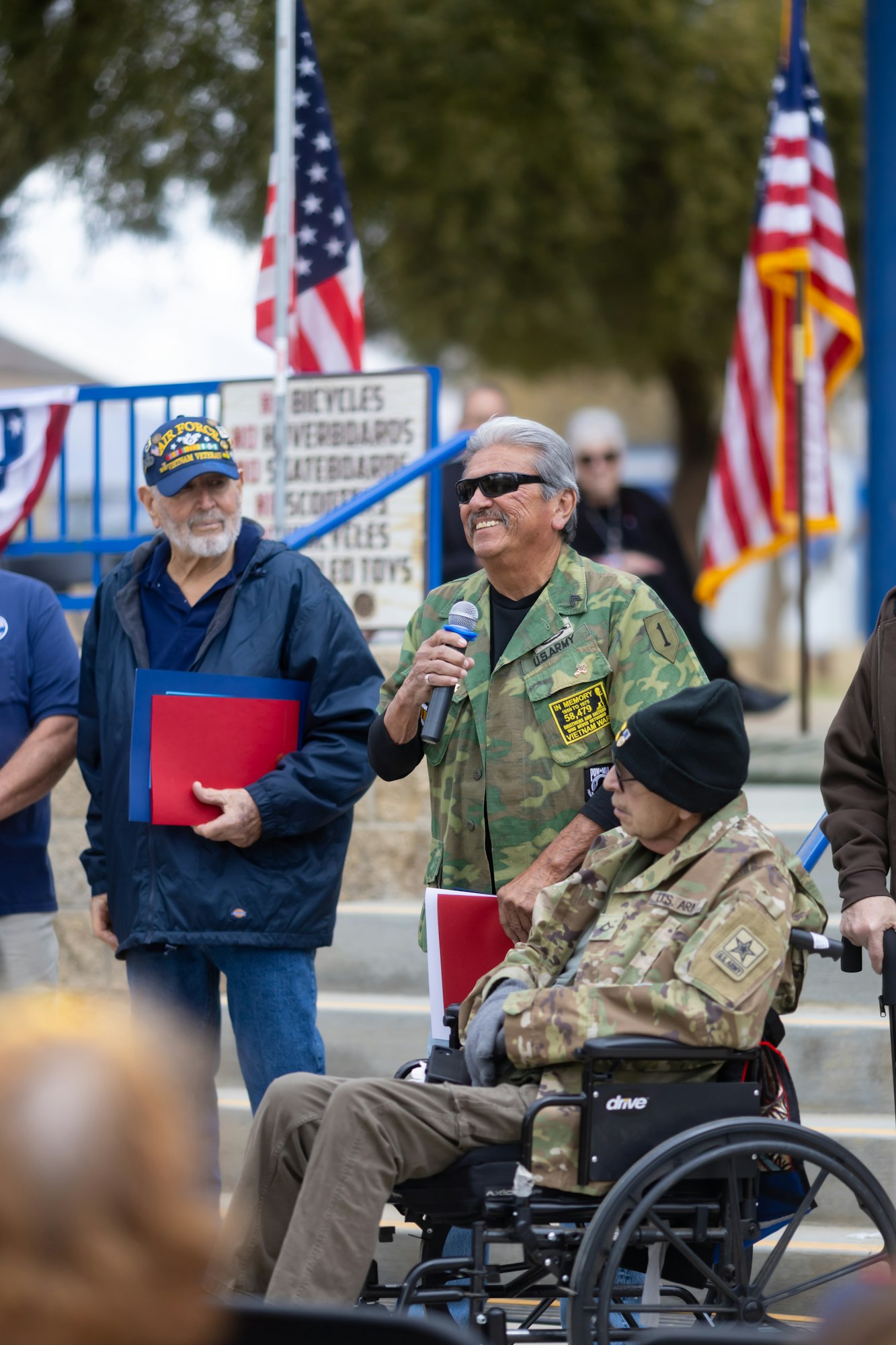 A veteran in a wheelchair with others standing, holding documents and a microphone, in front of American flags.