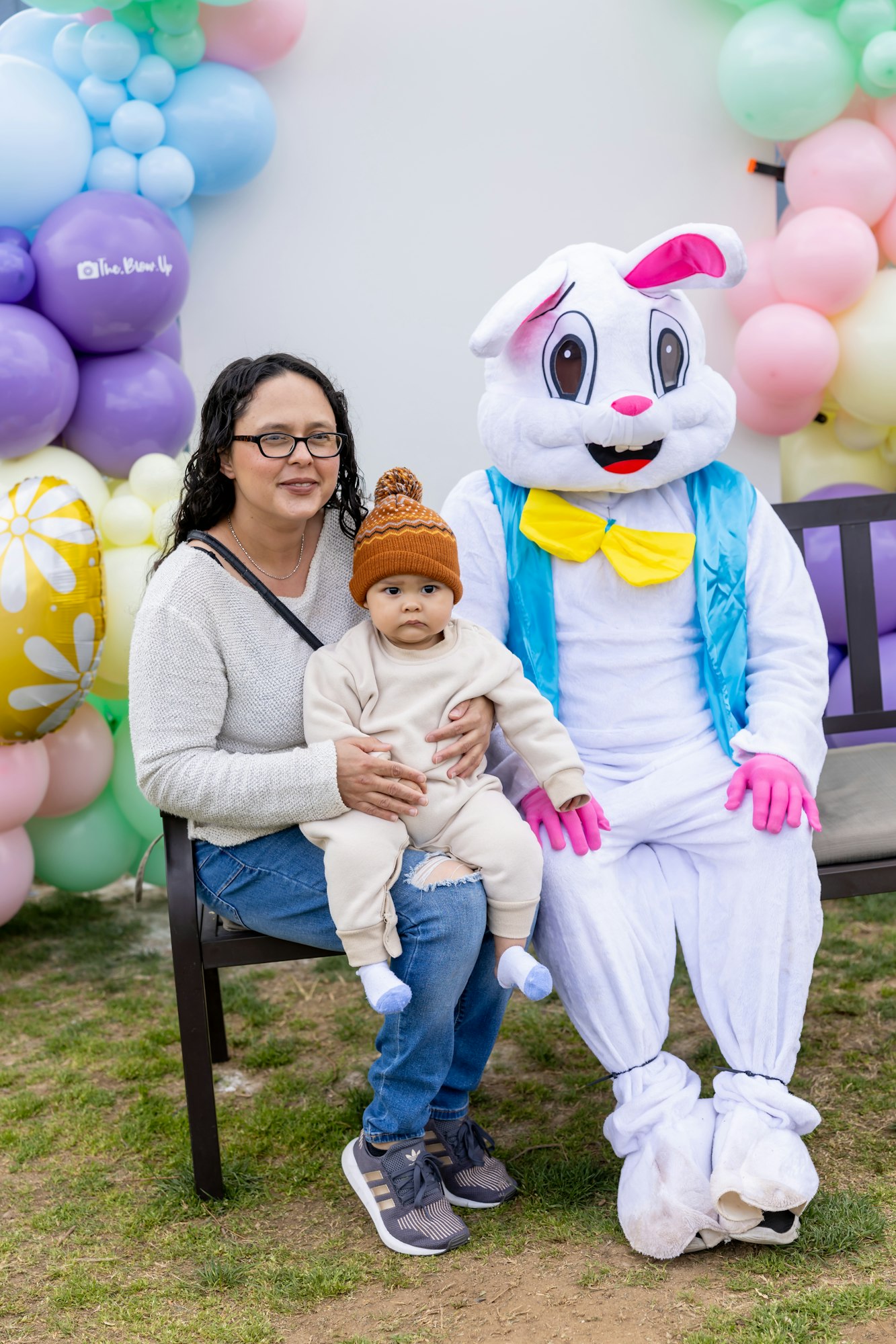 A woman and baby sitting next to a person in a bunny costume with colorful balloons in the background.