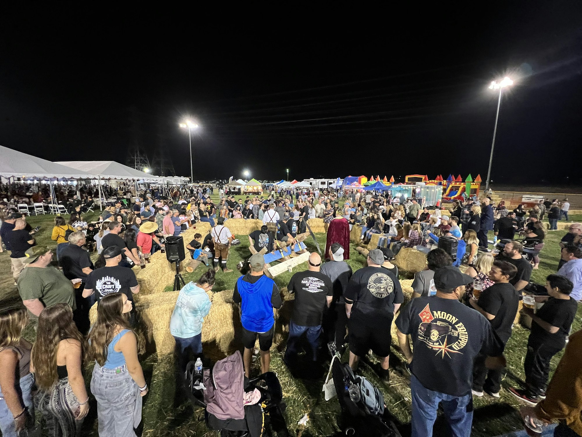 A nighttime outdoor event with a large crowd gathered around hay bales, tents, and attractions.