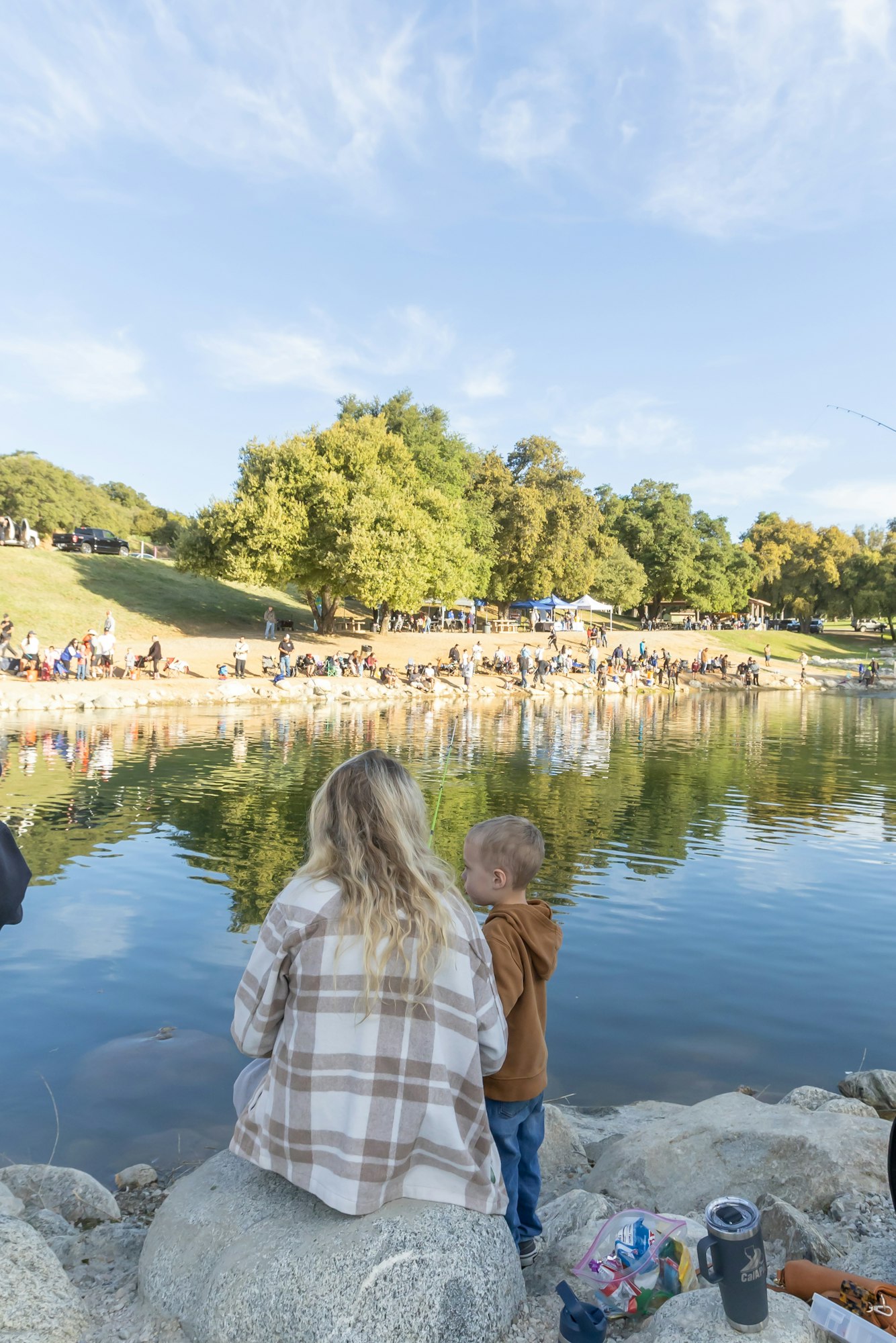 A person and child sitting by a lake with a fishing rod; people gathered on the opposite shore in a park setting.