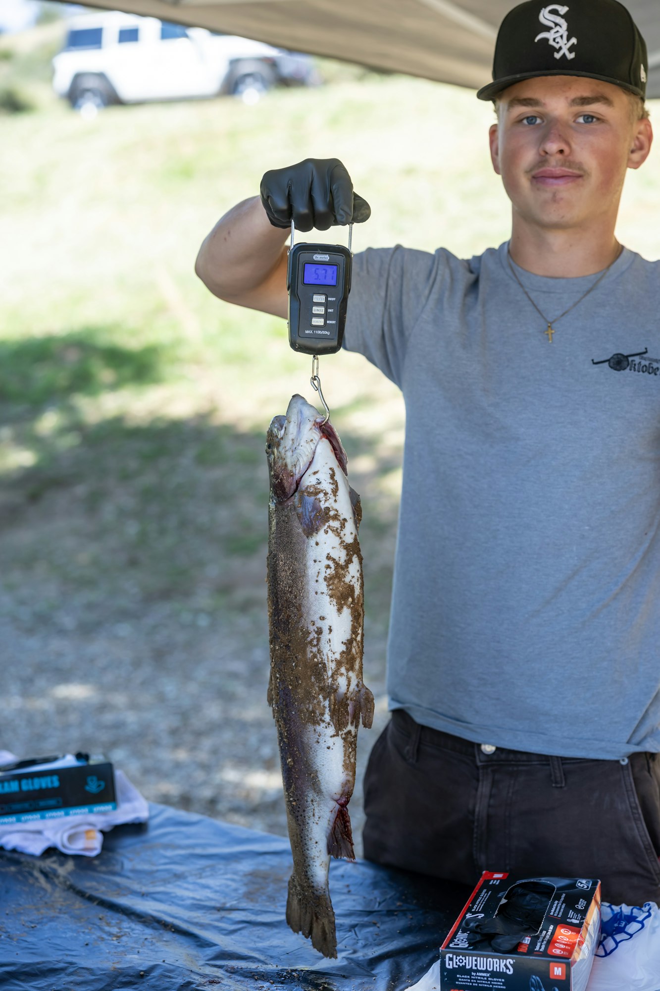 A person holds a large fish on a digital scale reading 5.7, wearing gloves with a car in the background.