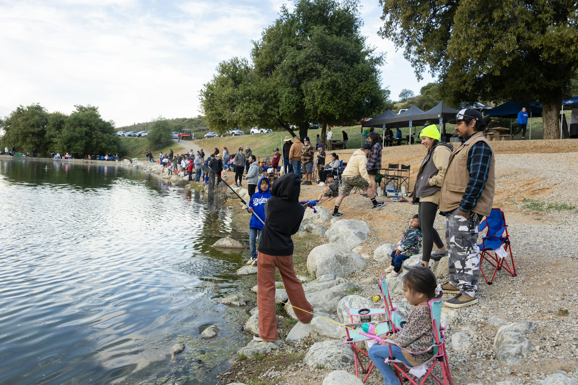 People fishing along a lakeside, with many families and children participating, enjoying a day outdoors near water.
