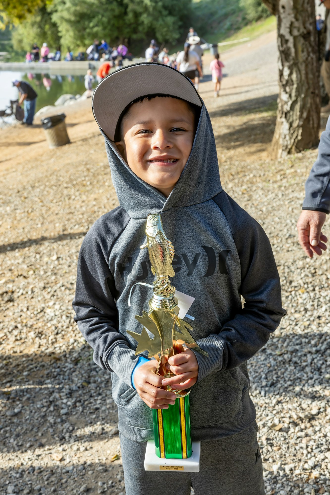 A smiling child in a hoodie holds a trophy with a fish figurine outdoors.