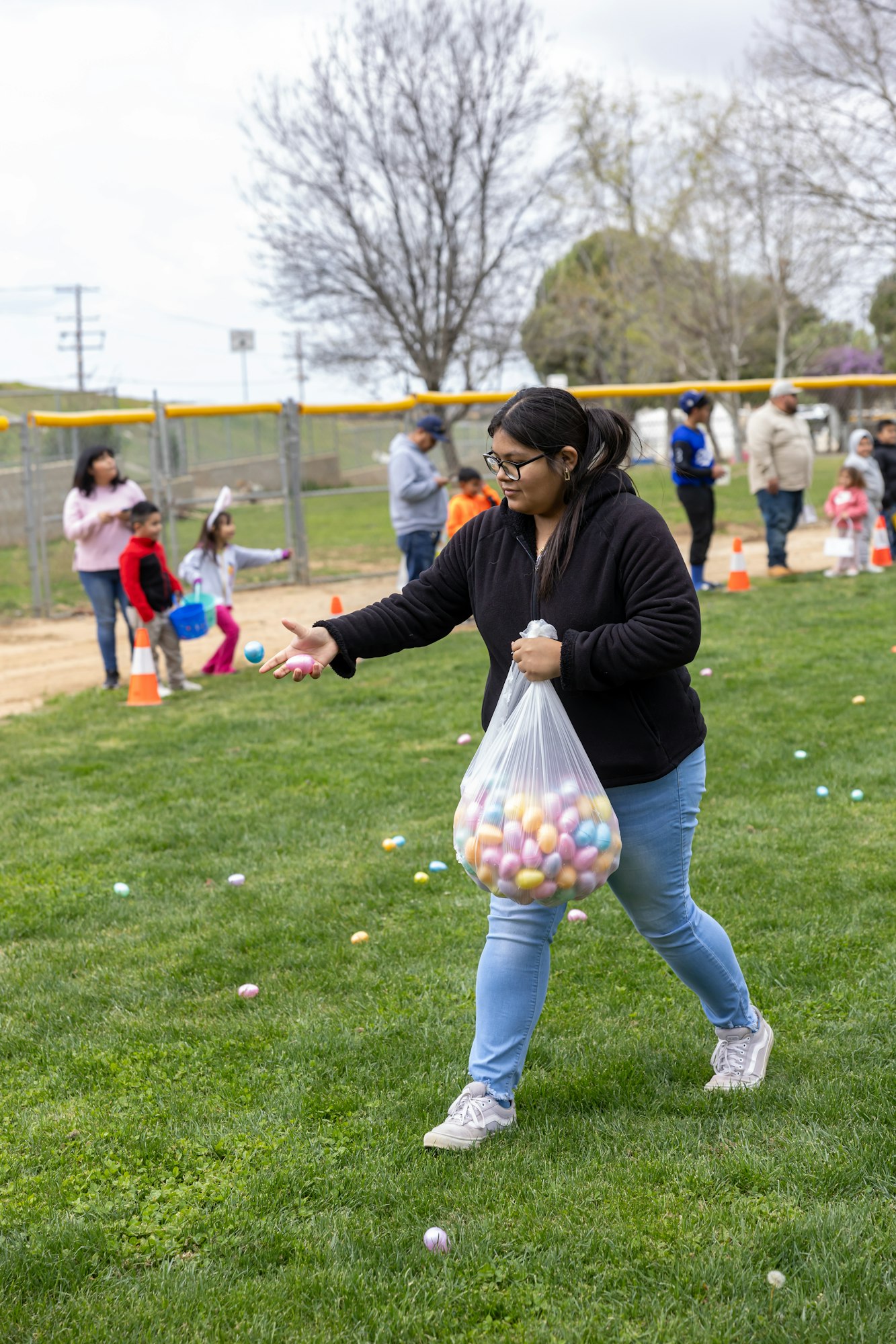 Woman placing colorful eggs on grass, children and adults nearby at an outdoor event.