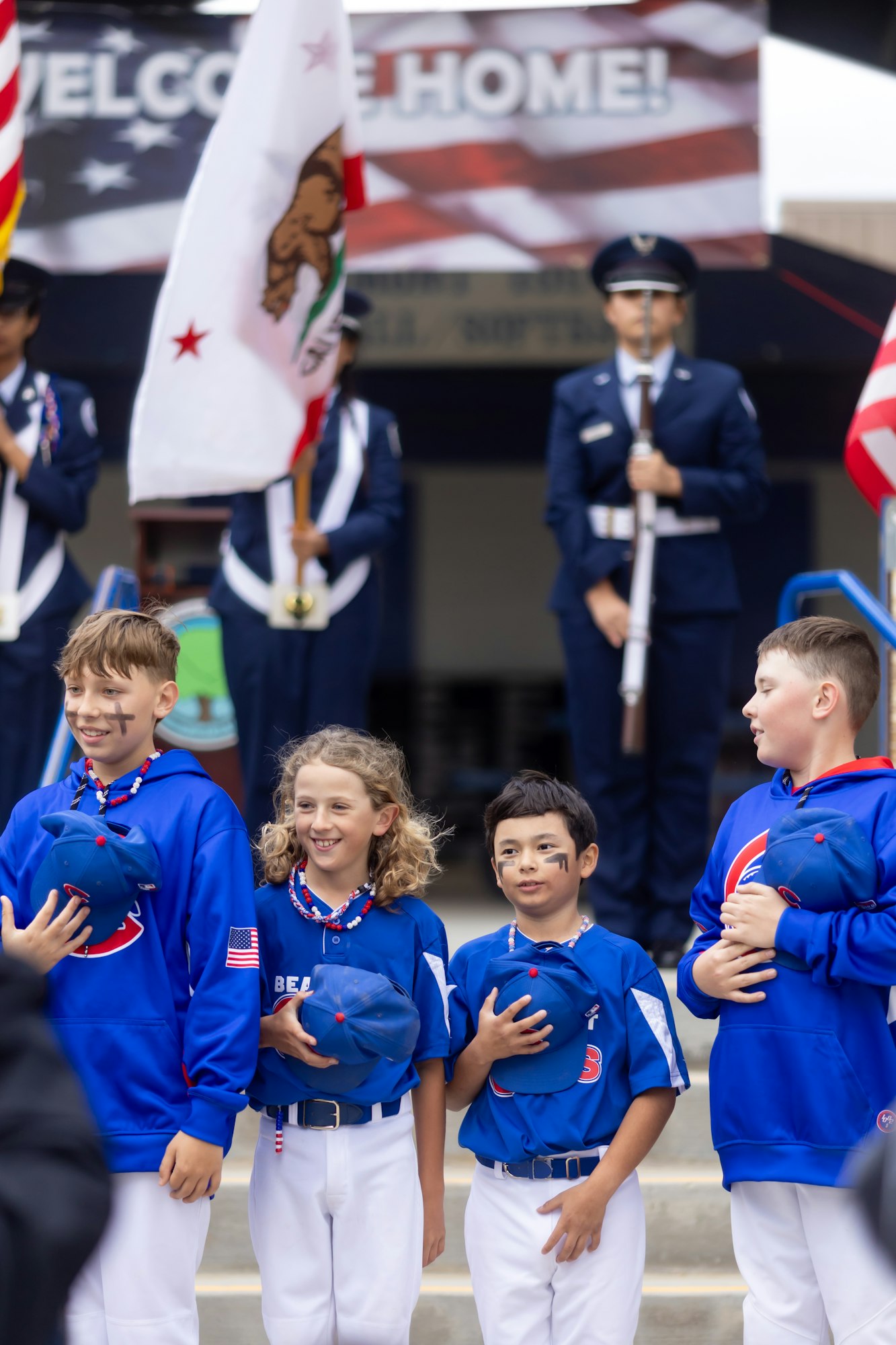A group of kids in sports uniforms holding hats, standing in front of a welcome home banner and flag bearers.