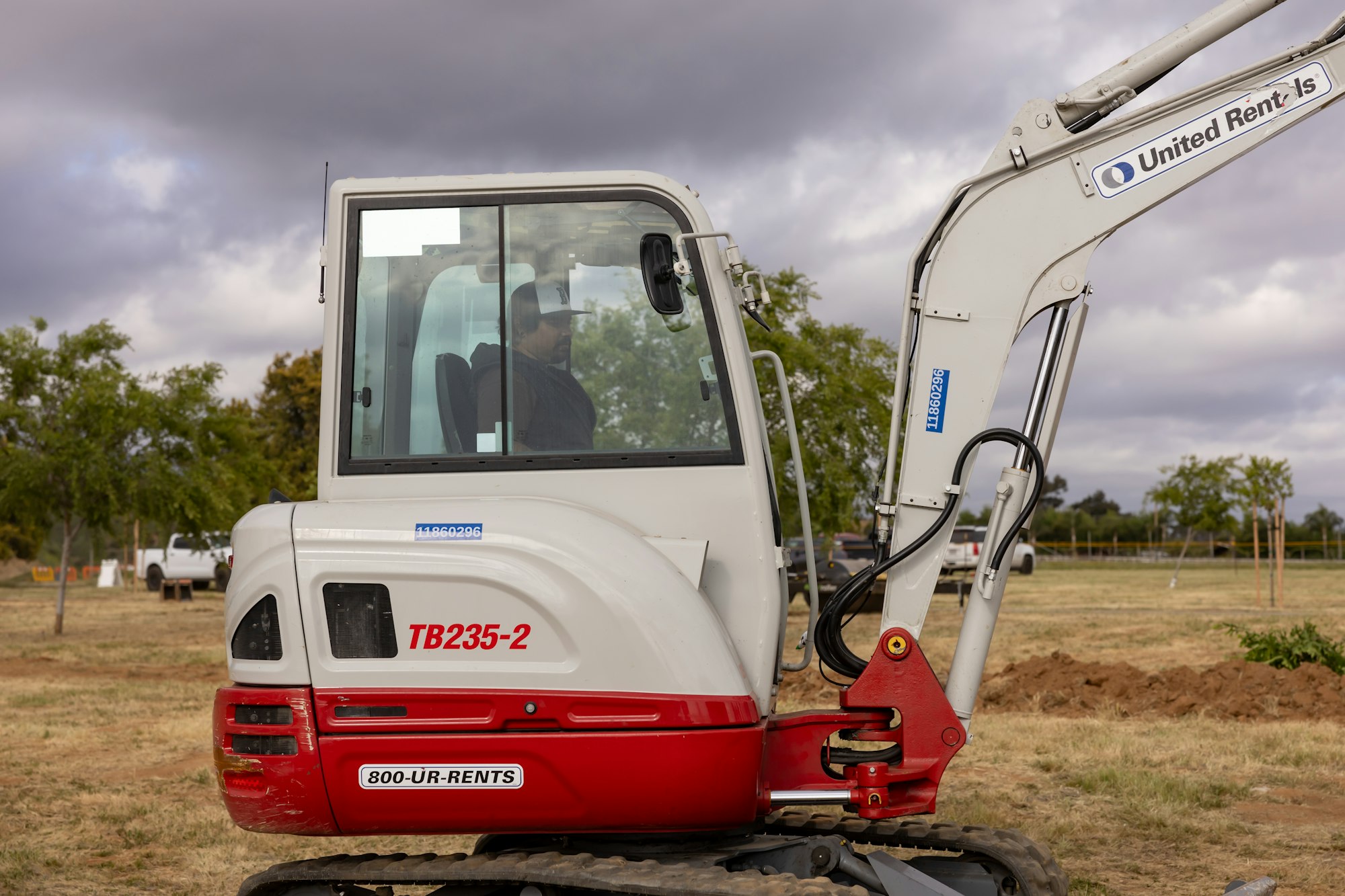 An excavator with “TB235-2” and “United Rentals” branding, on a grassy field under cloudy skies.