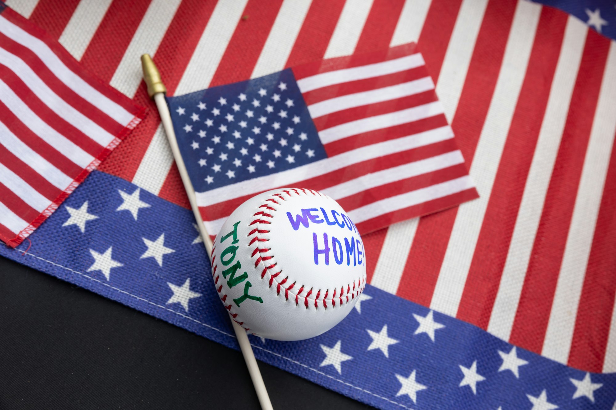 A baseball with "TONY" and "WELCOME HOME" on it, placed on an American flag with a small flag beside it.