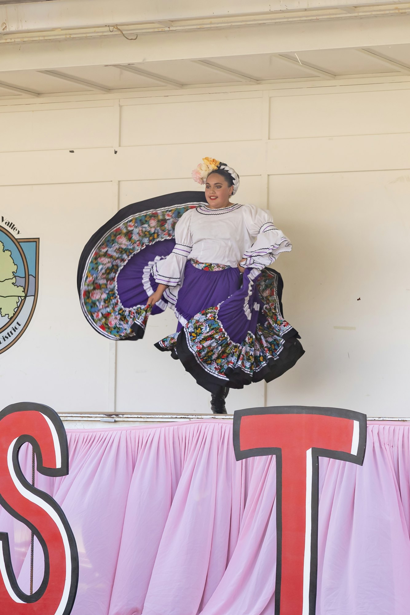 A dancer in traditional attire performs on stage, wearing a colorful dress with flowers in her hair.