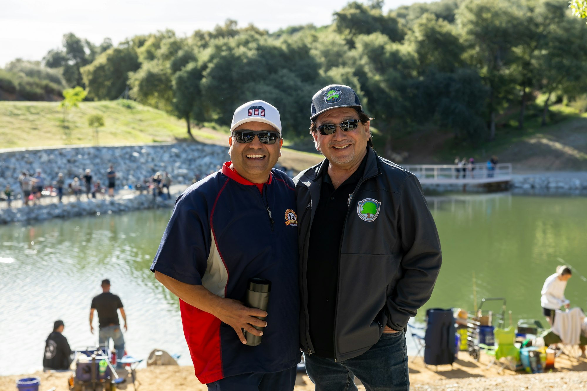 Two men smiling by a lakeside, wearing caps and sunglasses, with a scenic background of trees and water activities.