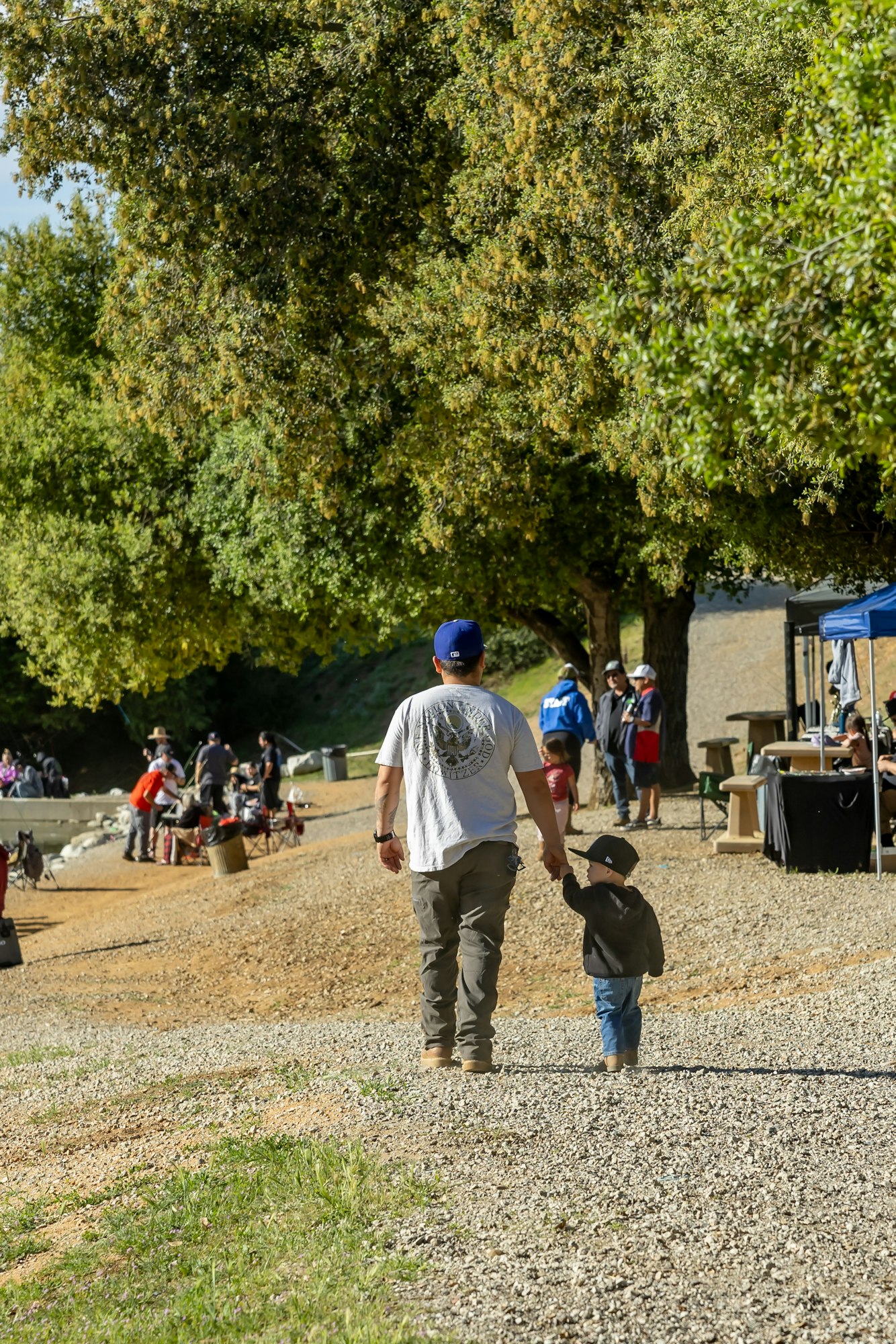 A man and child walk hand in hand on a gravel path under trees, with people and tents in the background.