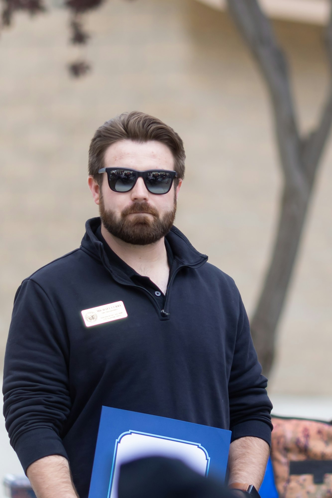 A person with sunglasses and a name tag, holding a blue-bordered certificate, stands outdoors.
