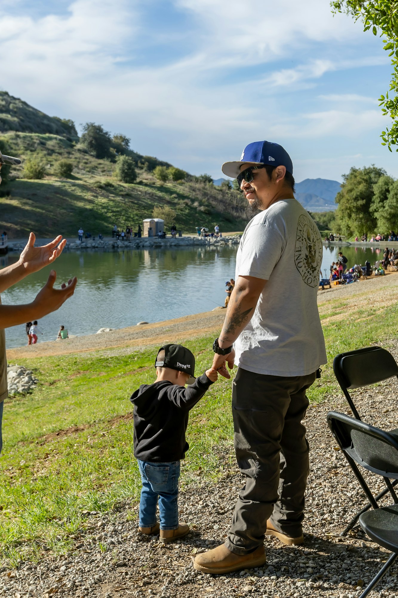 A man holding a child's hand near a lake; people in the background under a partly cloudy sky.
