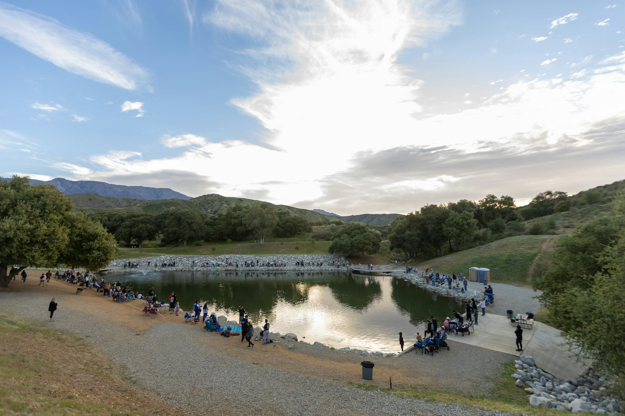 A scenic park with a pond, surrounded by hills, trees, and a crowd of people gathered along the water's edge.