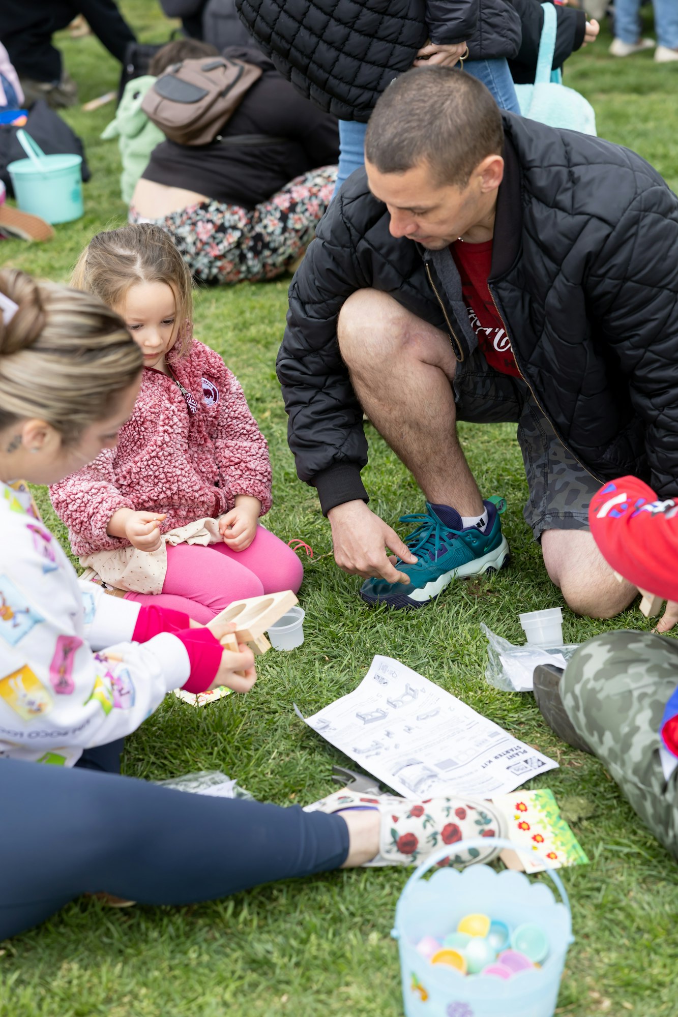 People sitting on grass, assembling a craft project with paper instructions and colorful eggs nearby.