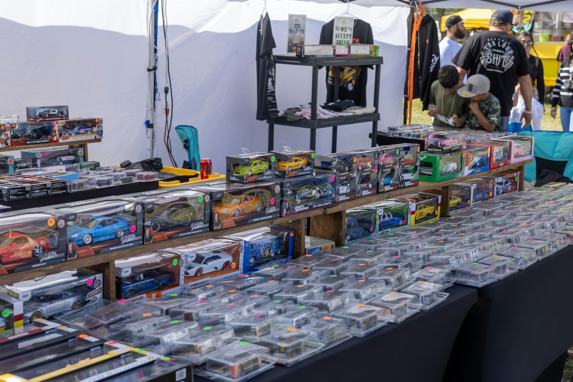 A market stall displaying model cars in boxes, with signs, merchandise, and people browsing.