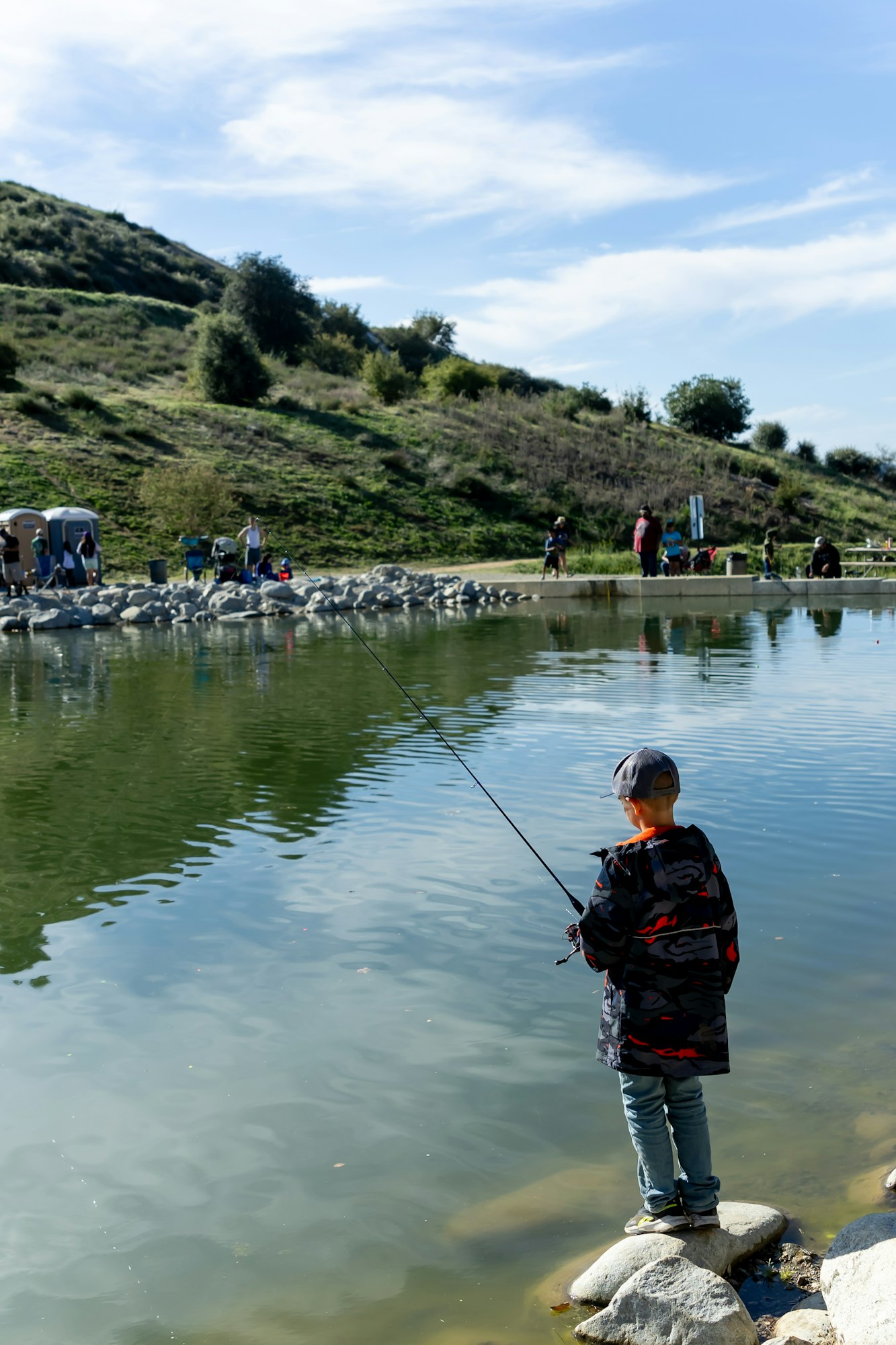 A child is fishing by a pond, surrounded by hills and people in the background.