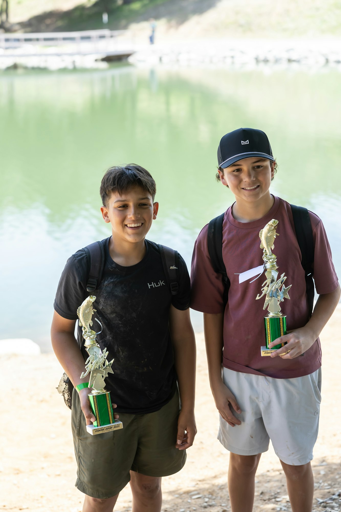 Two boys smiling, holding trophies by a lake.