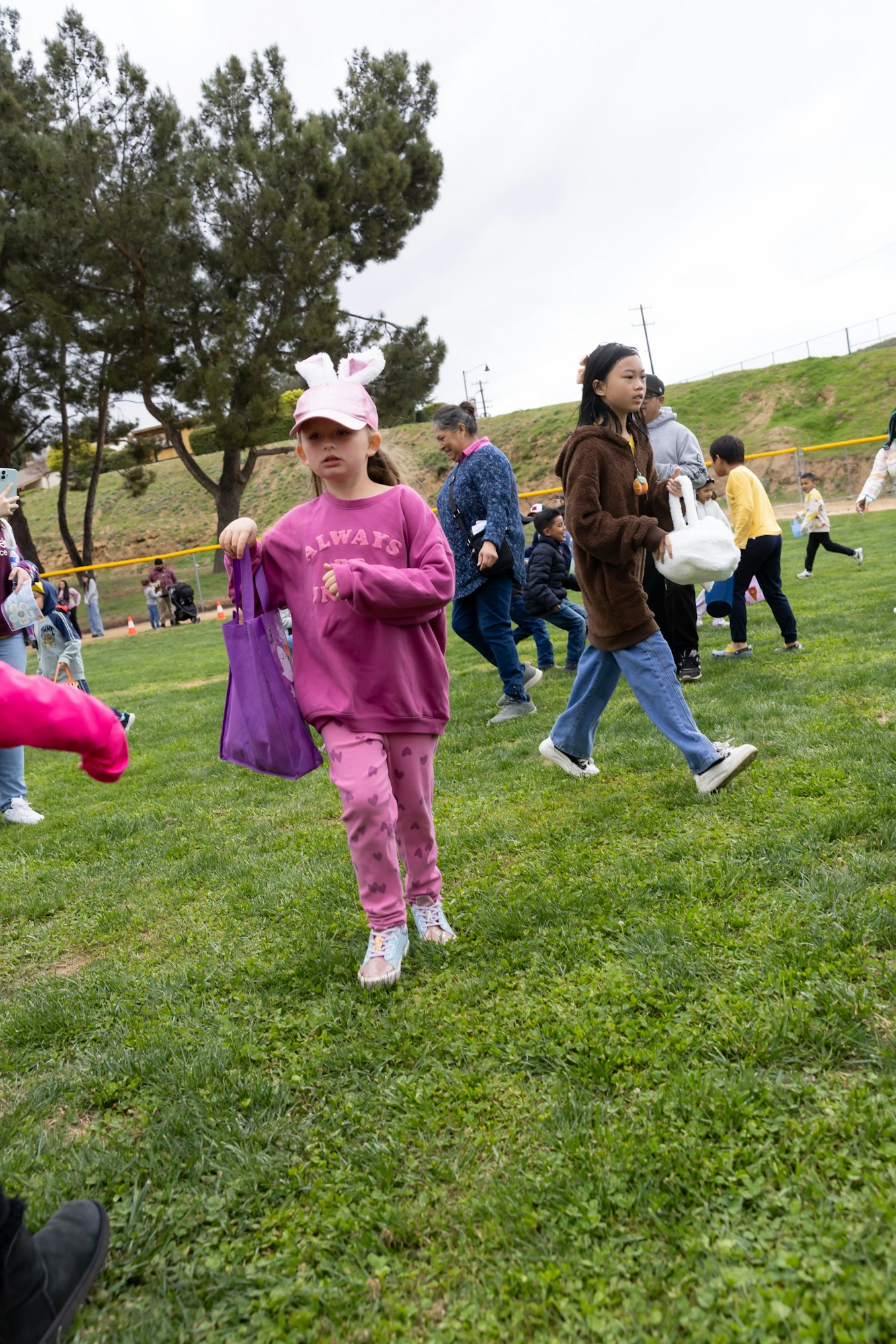Kids on a grassy field collecting items in bags, with trees and more people in the background.