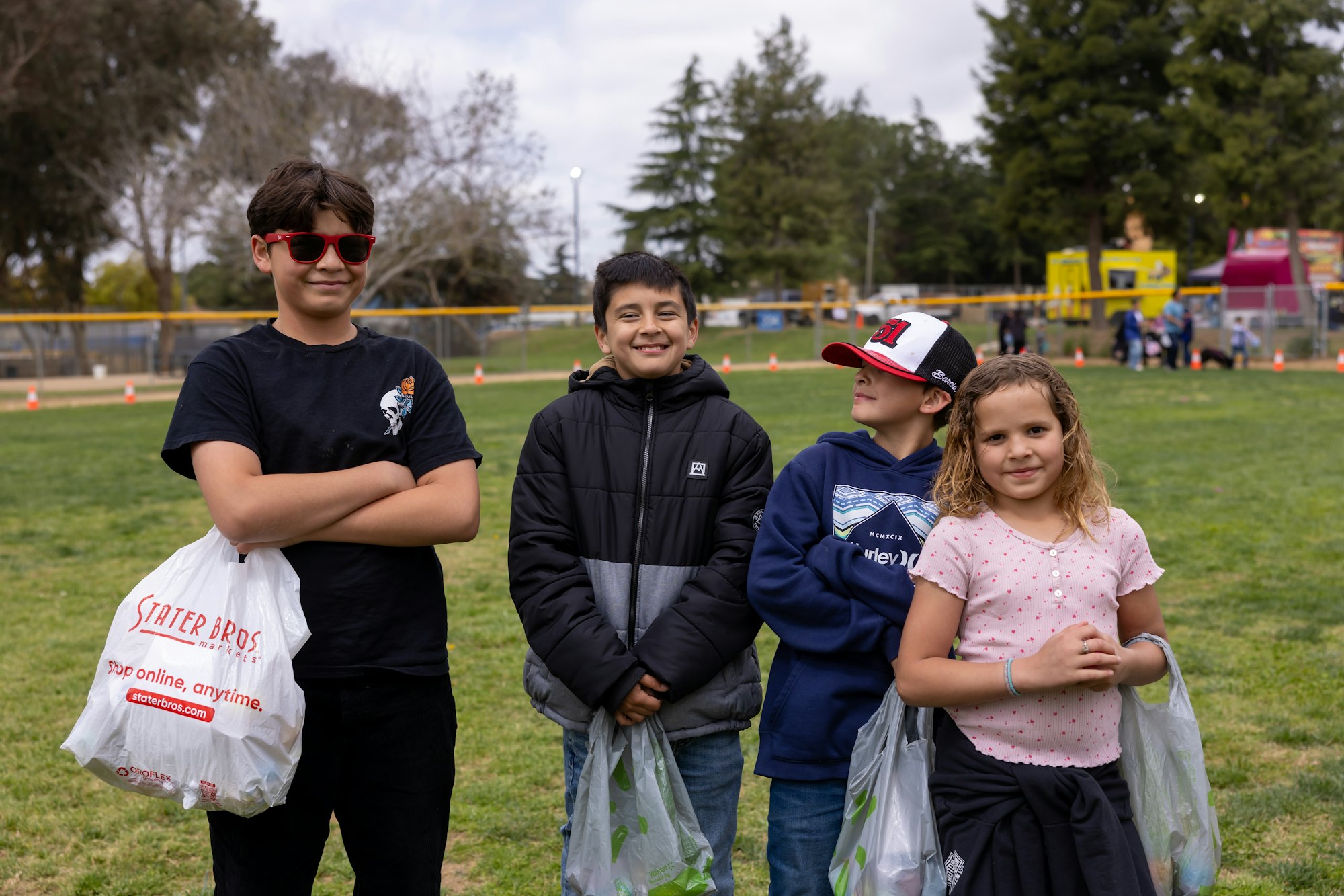 Four kids standing on grass, holding plastic bags, with trees and a playground in the background.