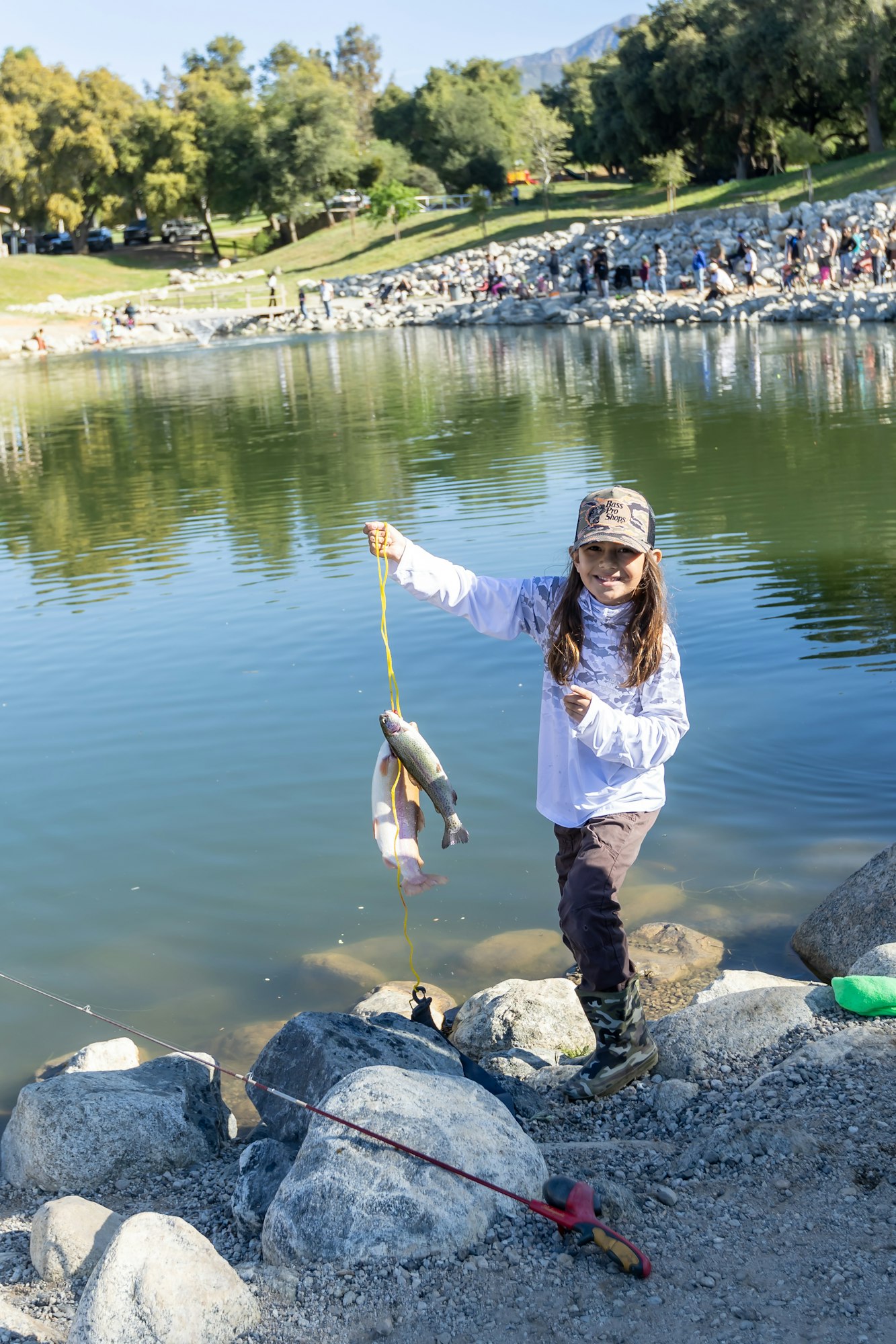 A child by a lake holds a string with two caught fish, wearing outdoor attire.