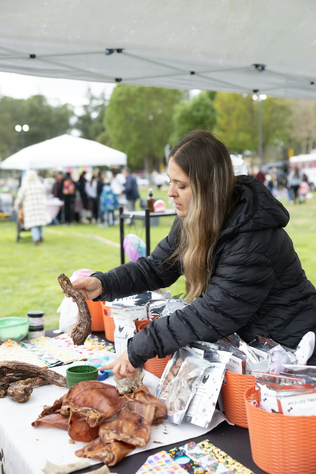 Woman in a black jacket at a market stall handling pet treats with customers in the background.