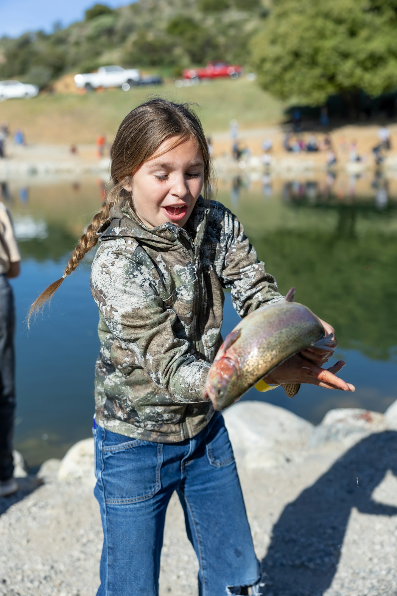A girl in a camo jacket holding a big fish by a lakeside, with a surprised expression.