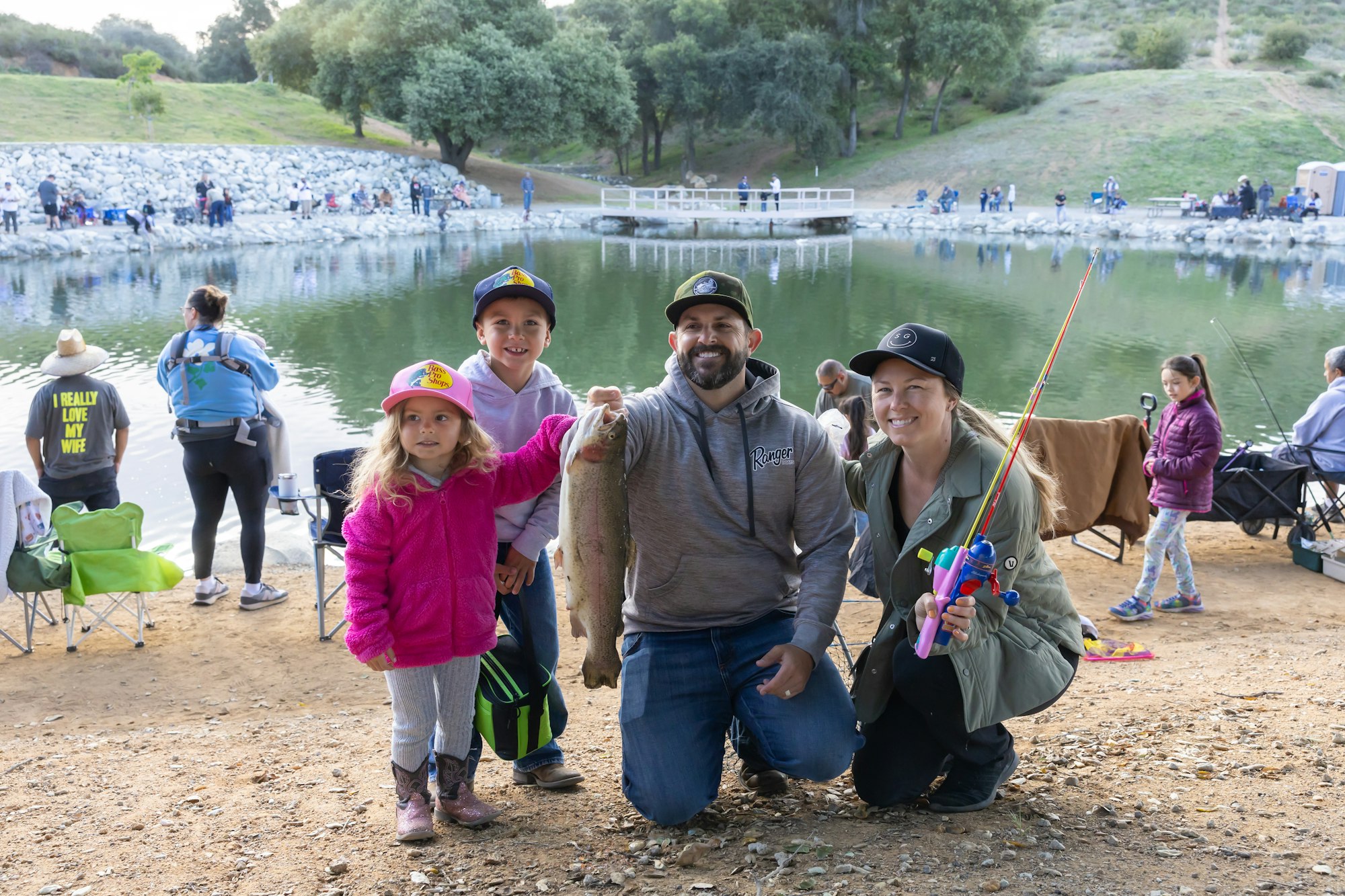 A family posing with a fish at a lakeside, surrounded by people fishing.