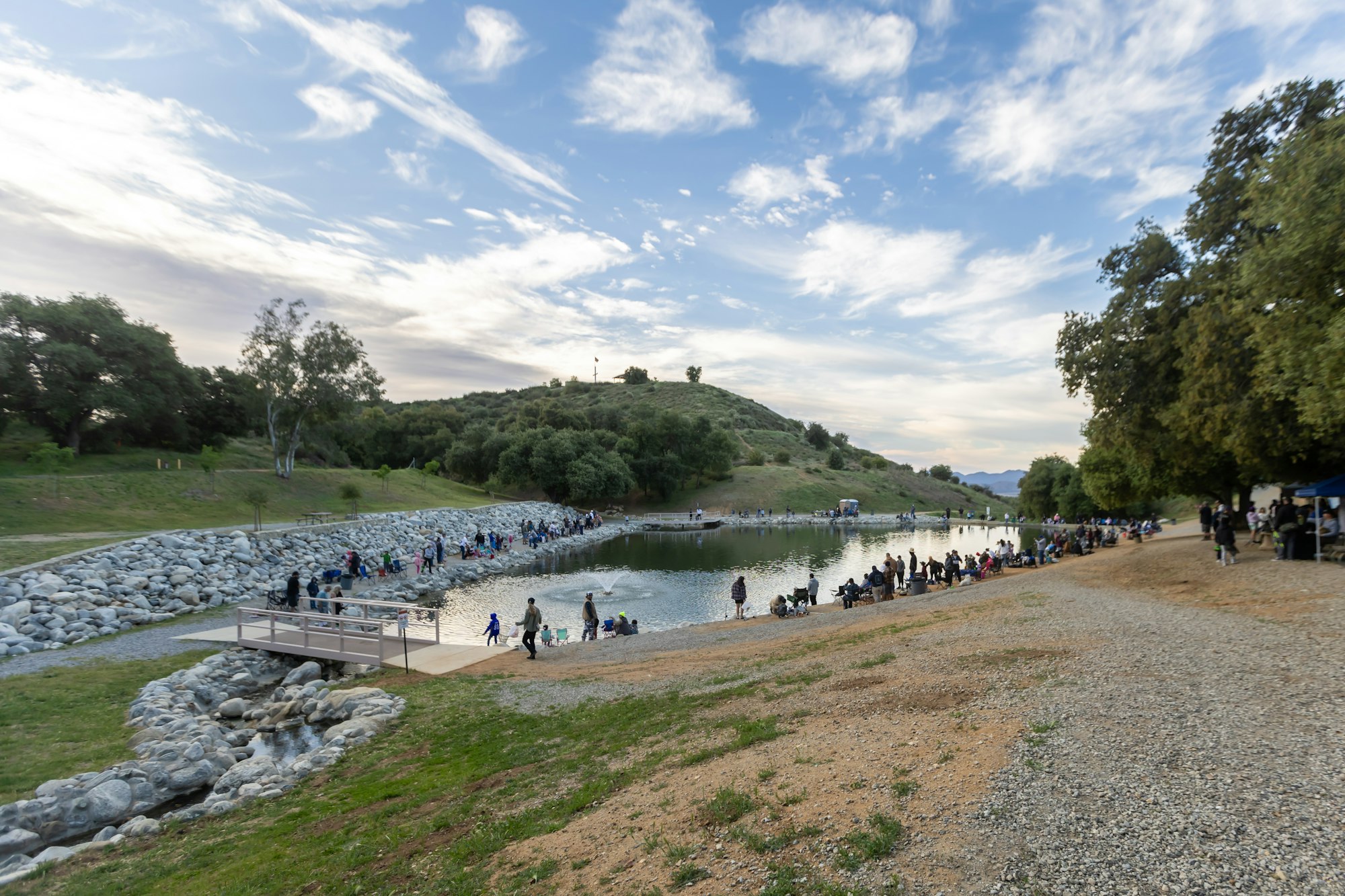 People gathered around a pond in a park-like setting under partly cloudy skies, engaging in activities.