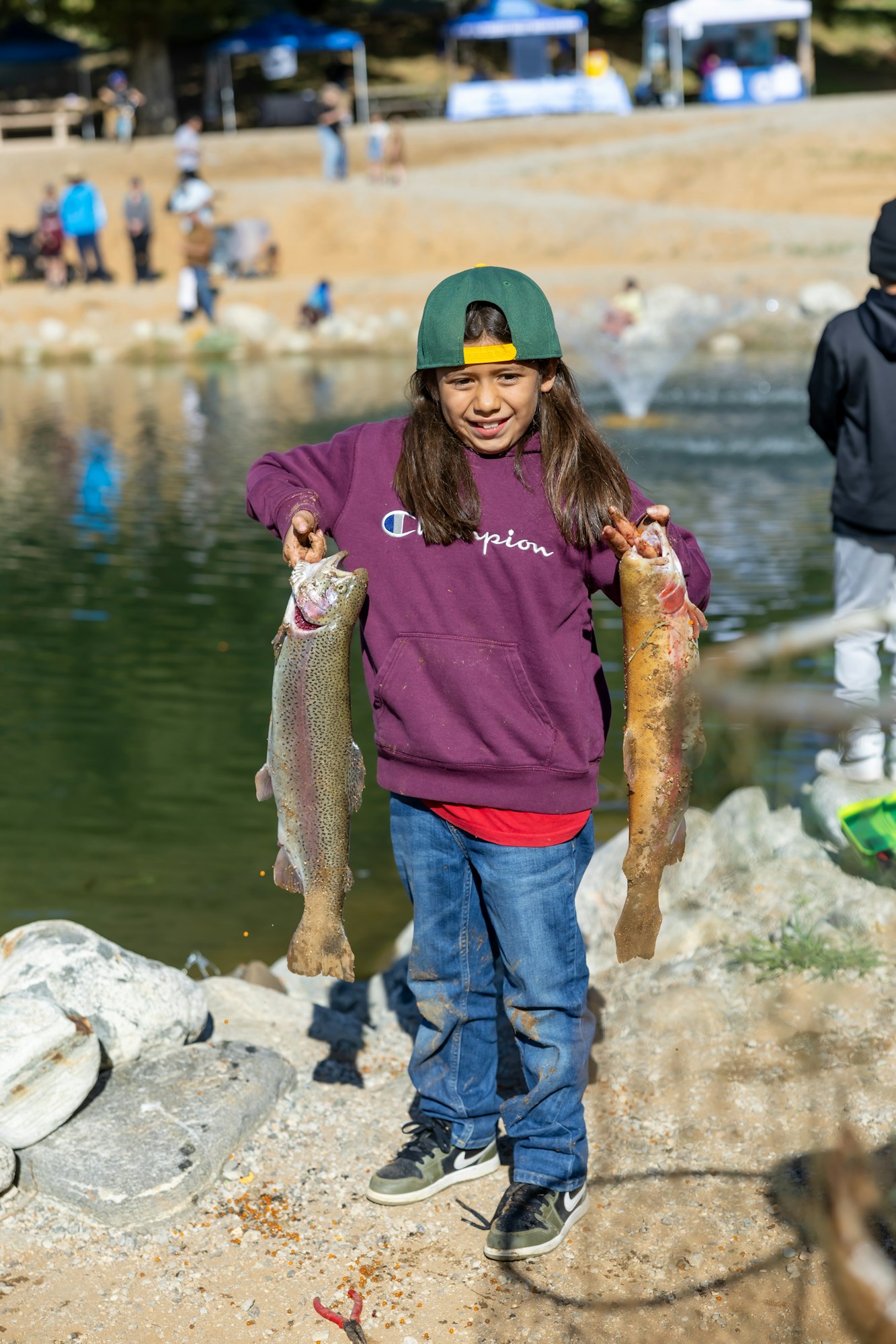 A child in a hoodie and cap holds two fish by a pond, with people and tents in the background.