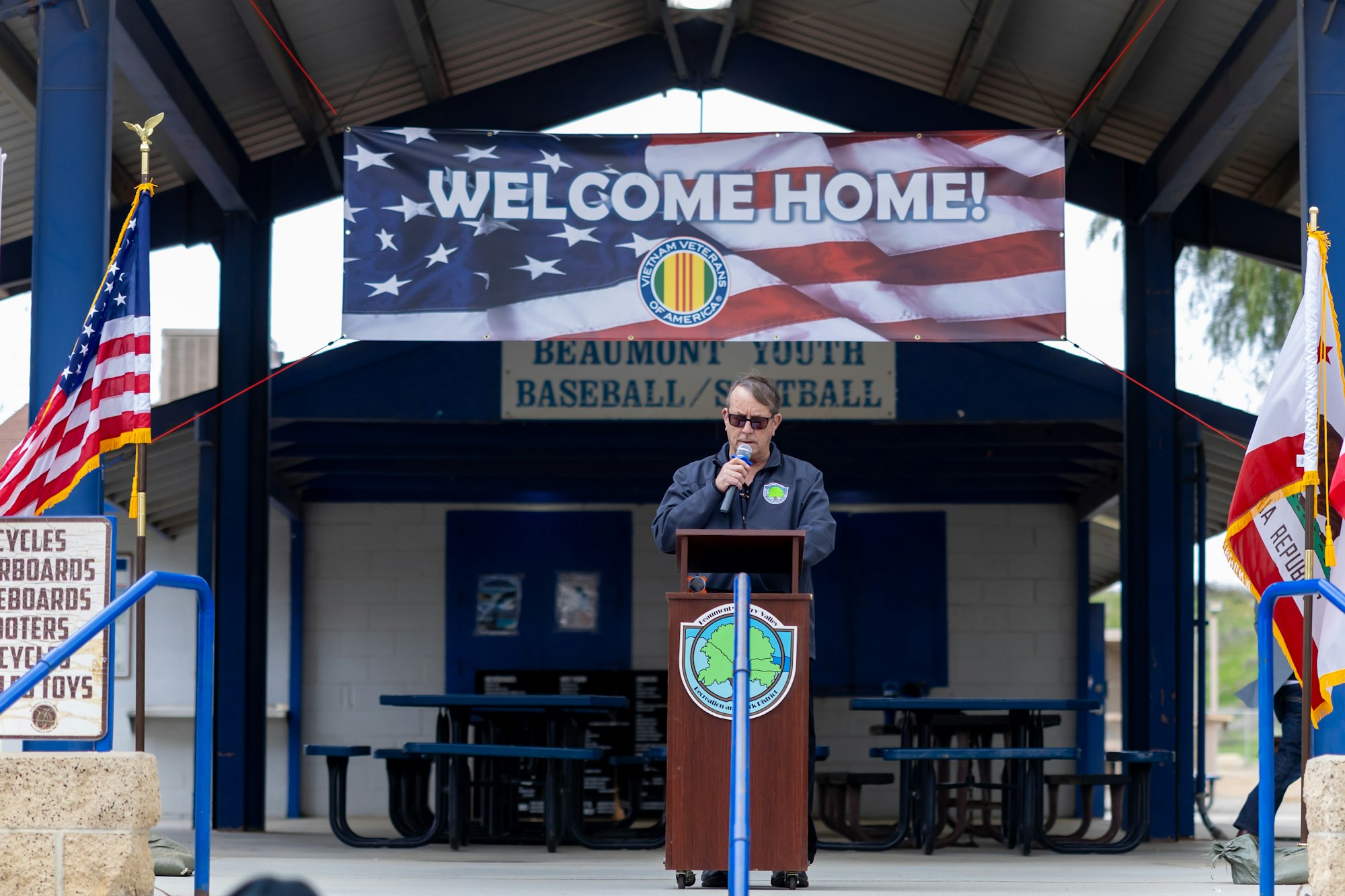 A man speaks at a podium under a "Welcome Home" sign with U.S. flag motifs, flanked by American and state flags.