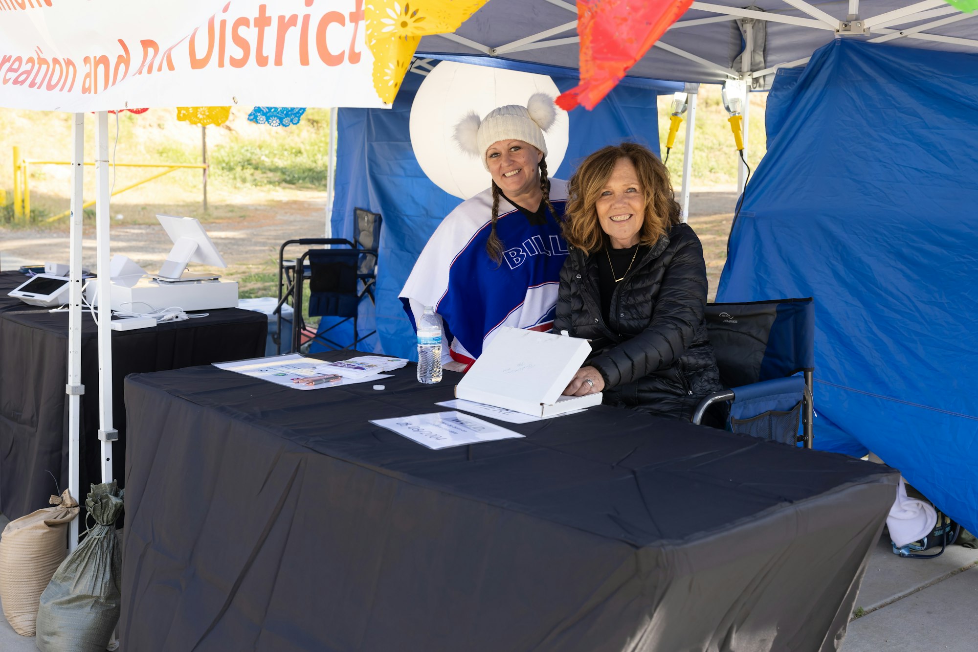 Two people at a booth under a tent, smiling at the camera. One is wearing a winter hat, and there's a "Recreation and District" banner above.