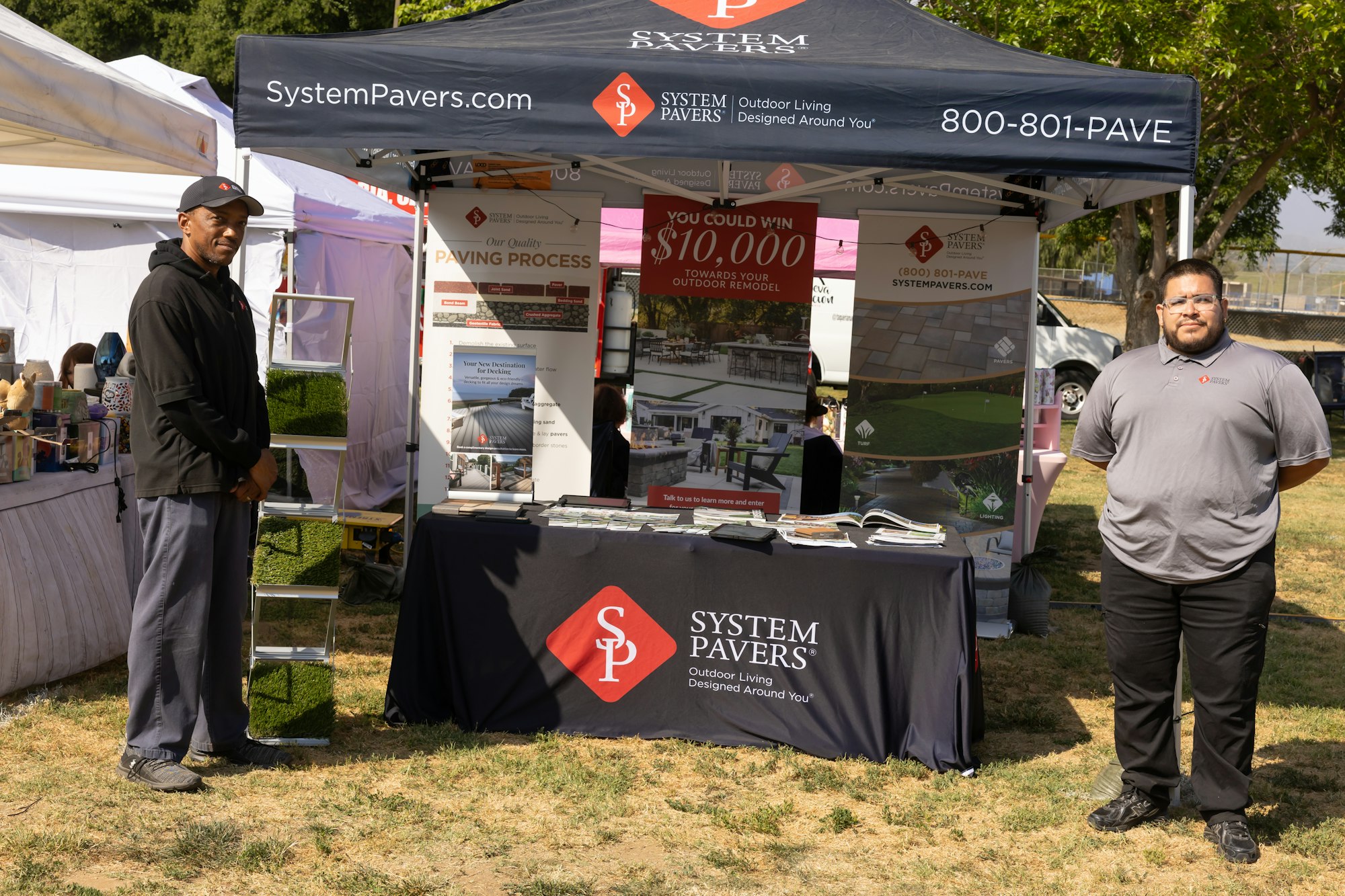 Two people stand by a System Pavers booth at an outdoor event displaying paving materials and promotional information.