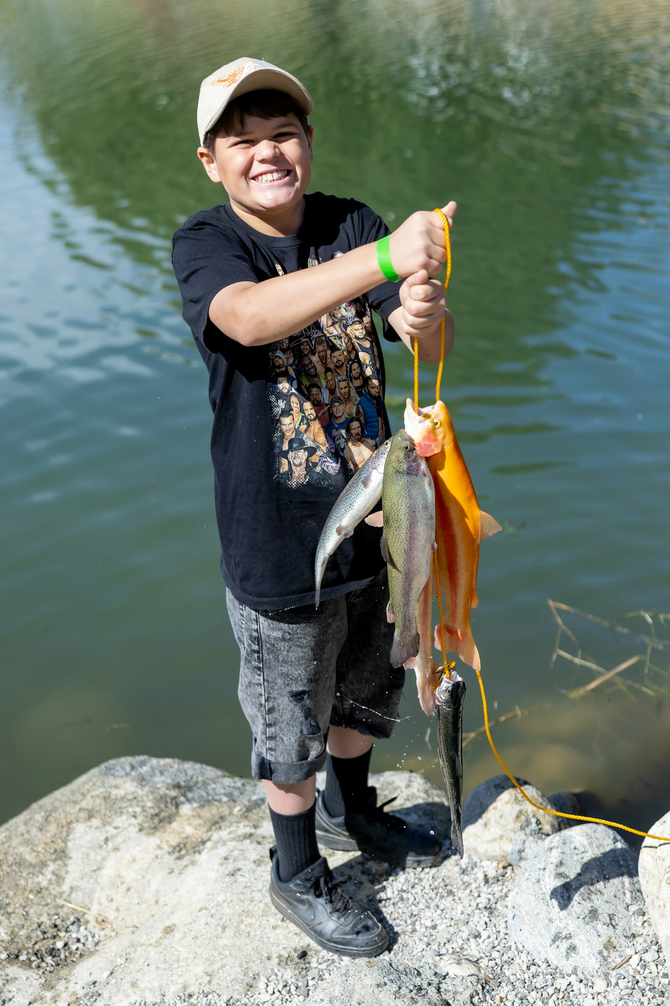 A smiling child holds up several fish by a lake, standing on a rocky edge.