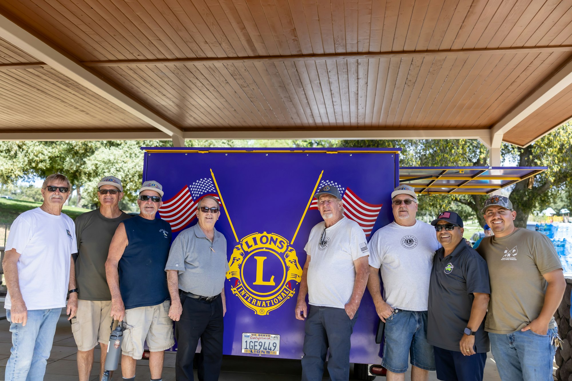 A group of people standing in front of a trailer with a Lions International logo and American flags, under a wooden gazebo.