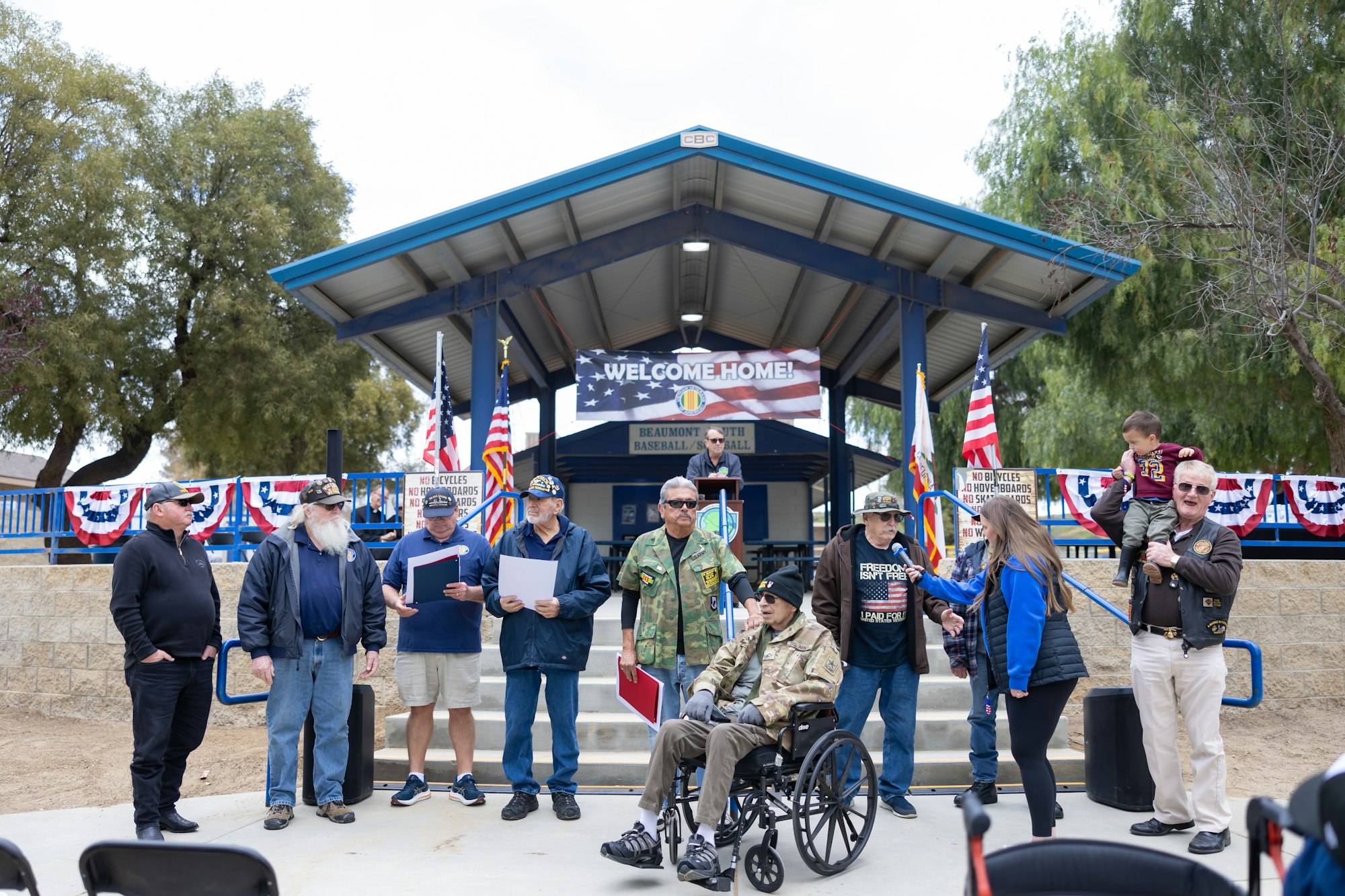 A group of people, some in military attire, are gathered with American flags and a "Welcome Home" banner at an outdoor event.