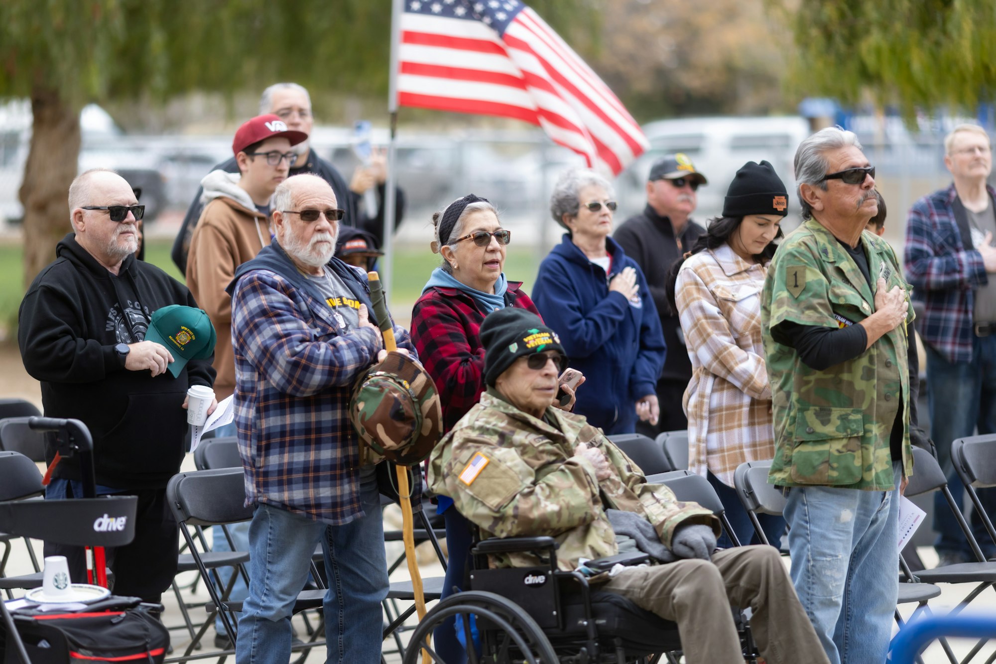 A group of people, some in military attire, stand with hands over hearts, near an American flag.