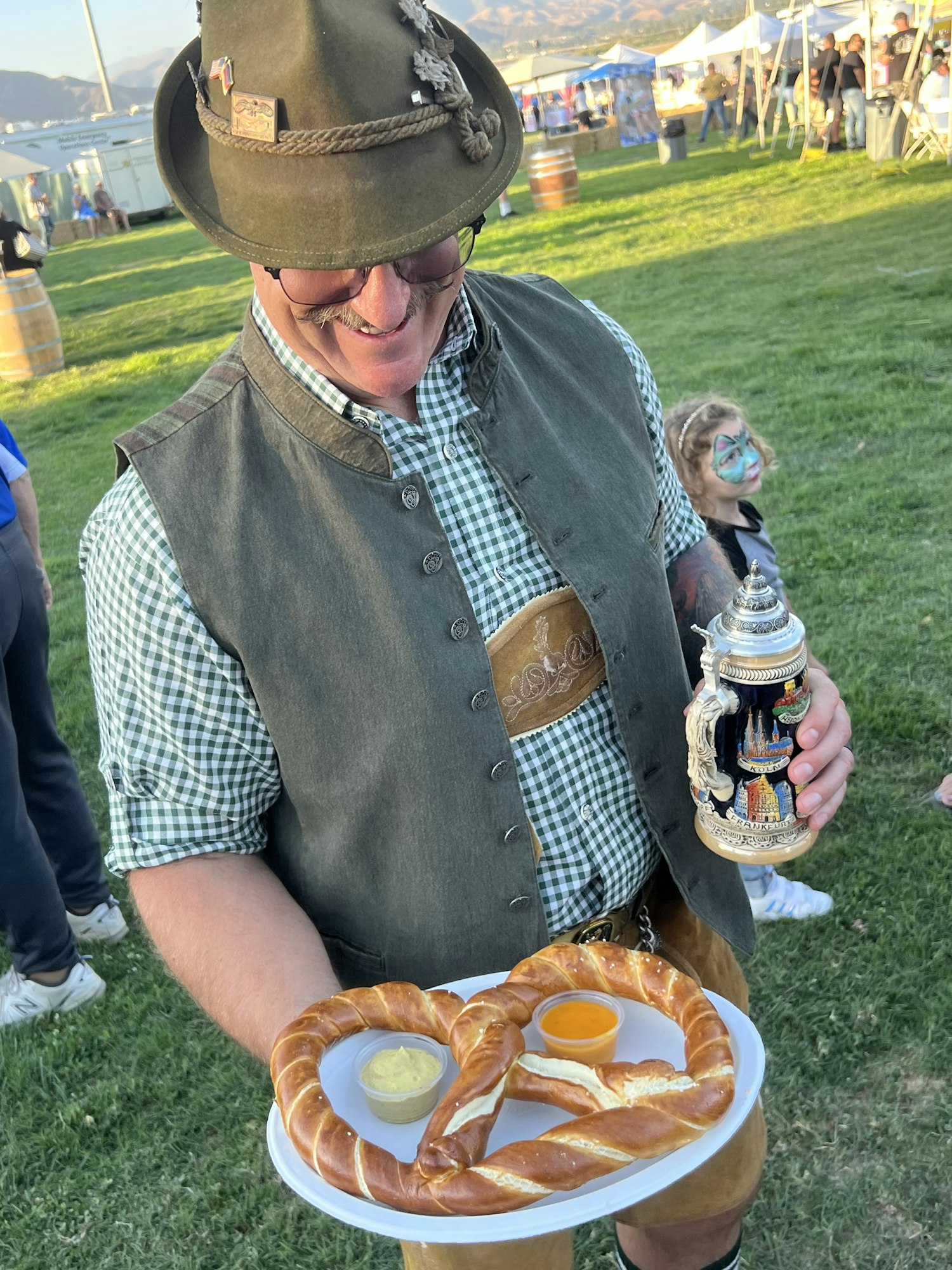 Man in traditional attire with a large pretzel and sauces on a plate, holding a beer stein at an outdoor event.