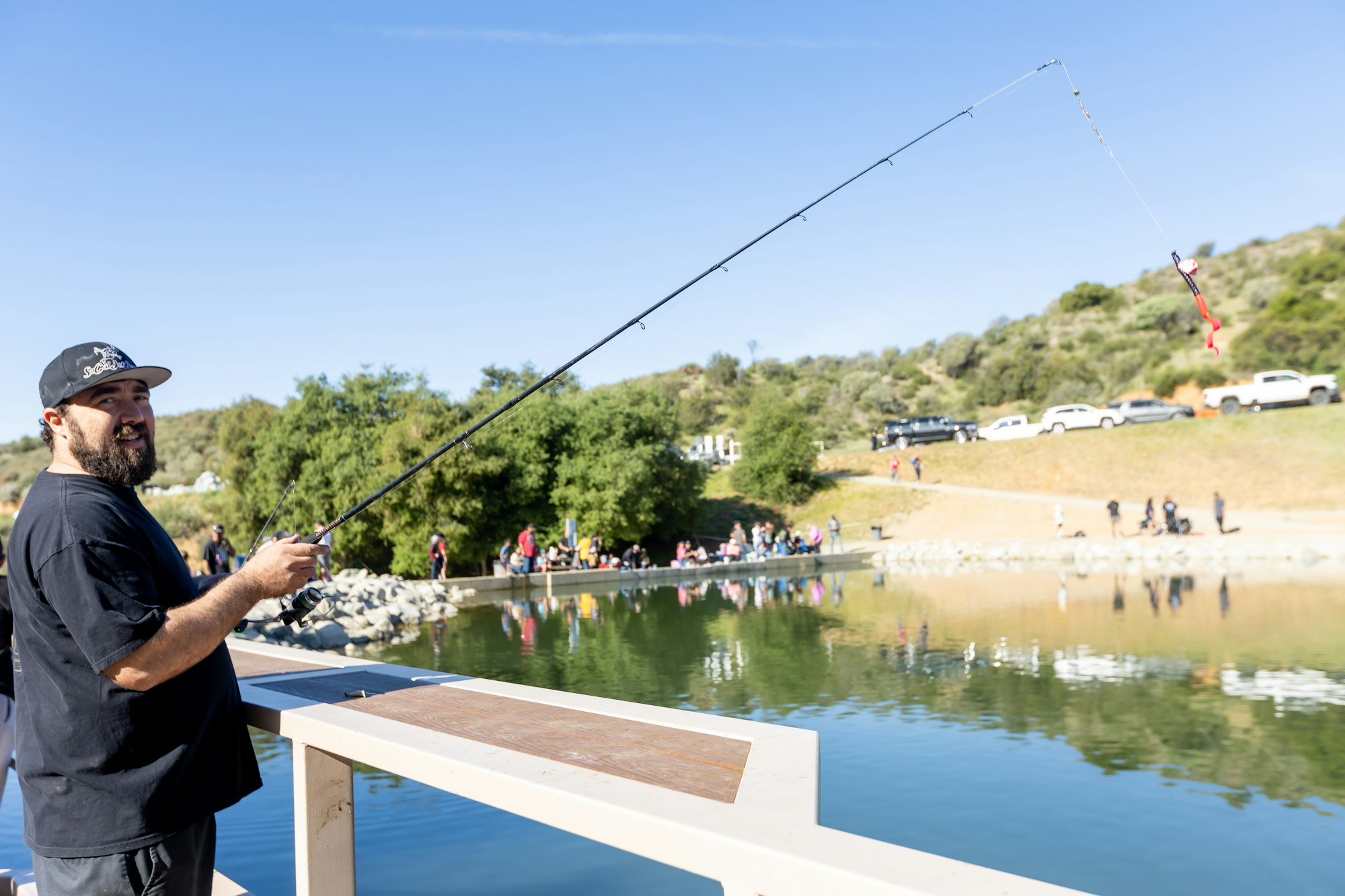 A person fishing by a pond with others in the background, under a clear blue sky.