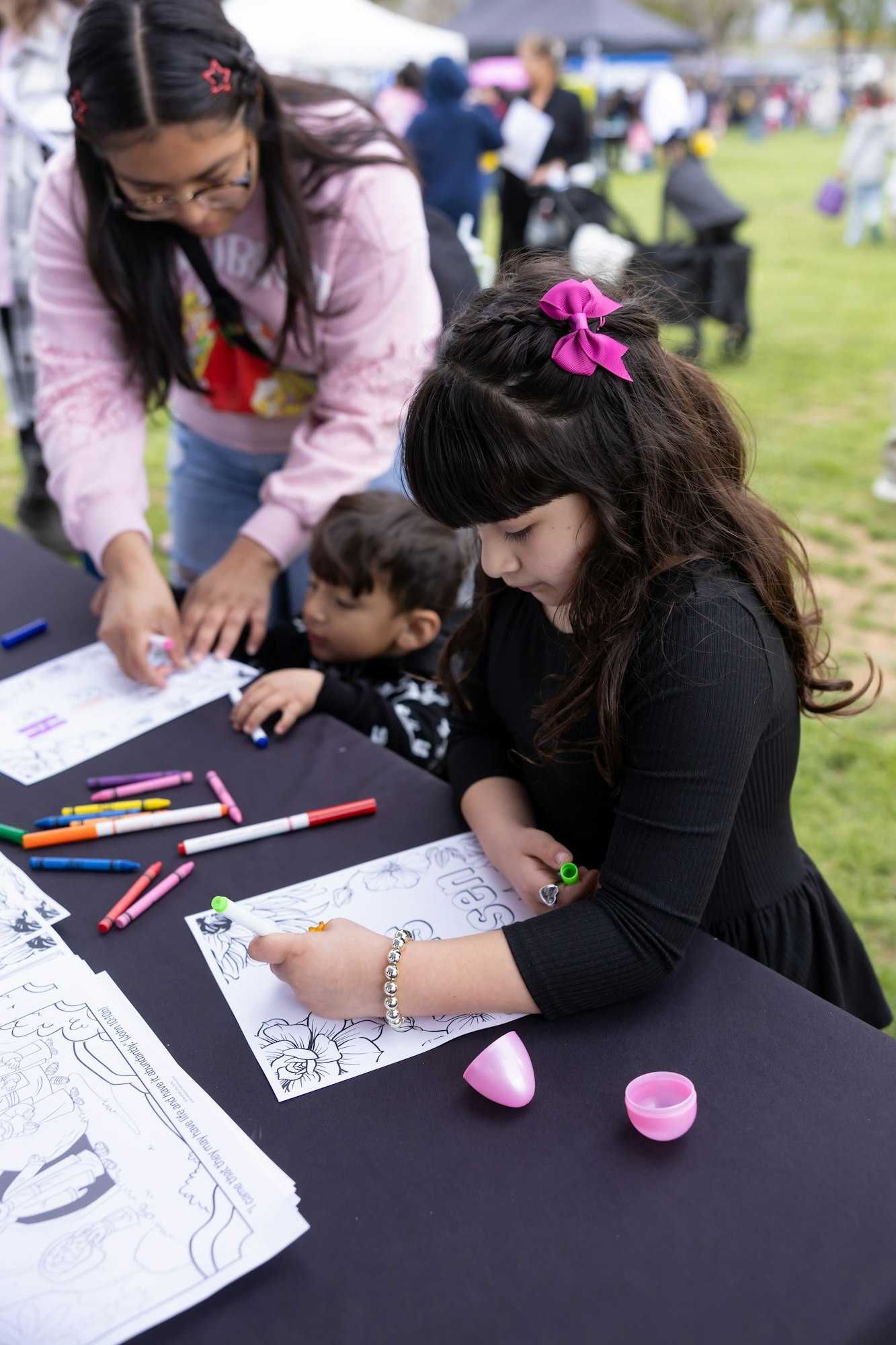 A child colors a sheet with markers at a table, supervised by an adult. Other people are in the background at an outdoor event.