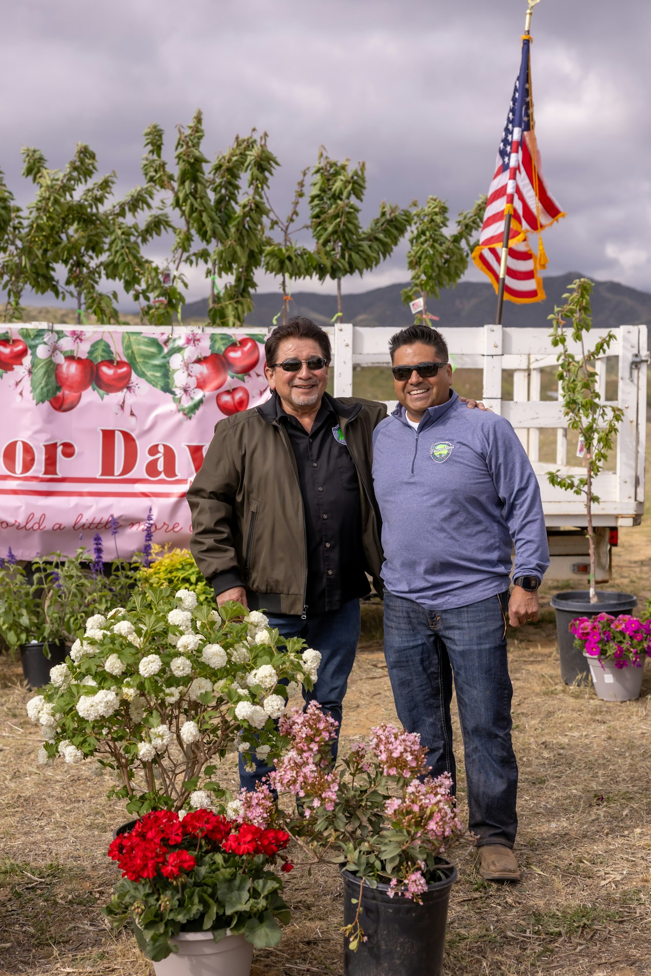 Two people smiling at an outdoor event with flowers and a banner. An American flag is visible in the background.
