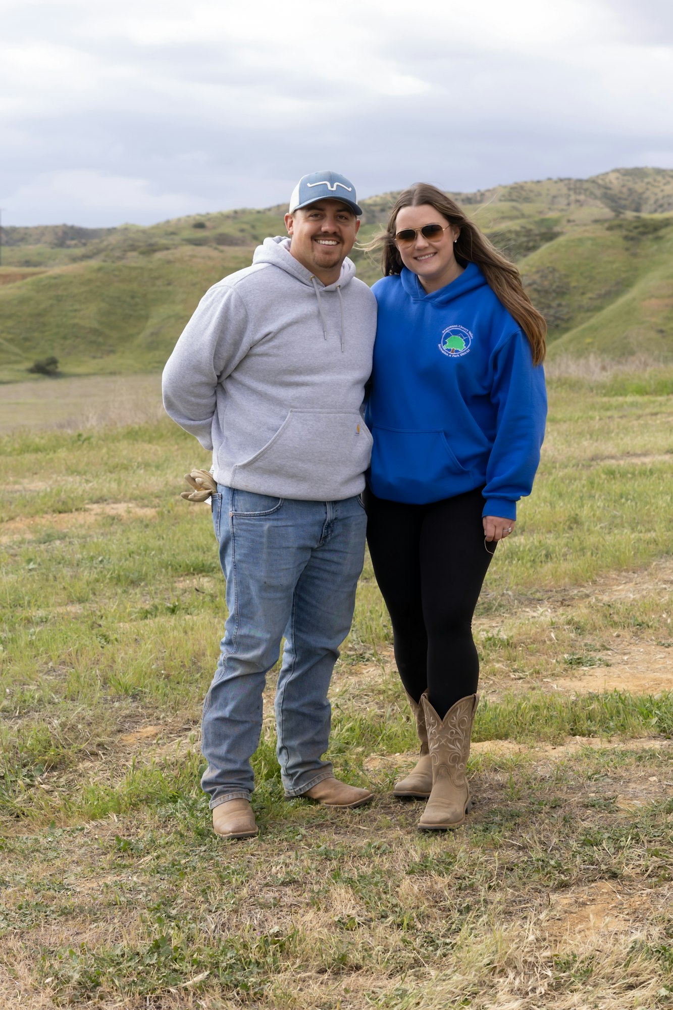A man and woman in casual attire standing on a grassy hill with a scenic landscape backdrop.