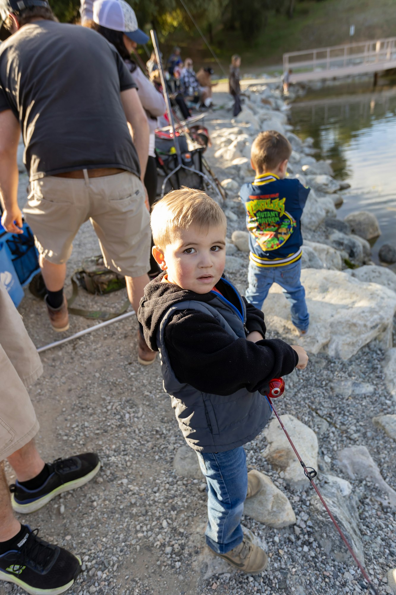 Children and adults fishing by a lakeside, standing on rocks and gravel. A boy holding a fishing pole looks toward the camera.