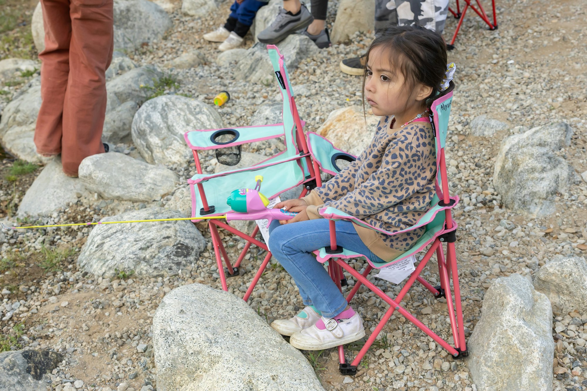 A child sits on a folding chair with a toy fishing rod, surrounded by rocks and other people's legs.