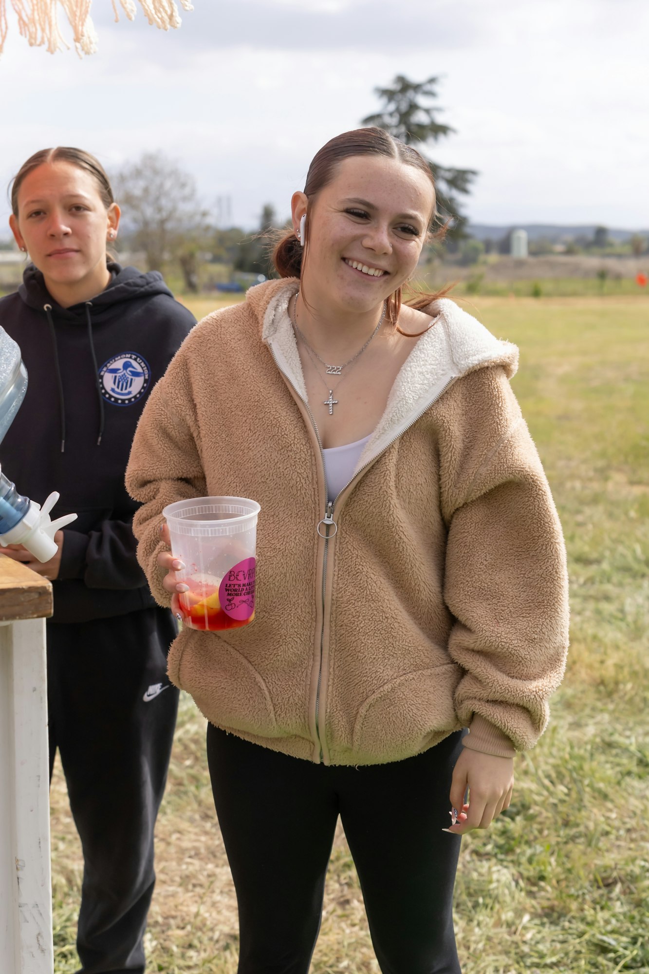 Two people standing outdoors; one is smiling, holding a drink, and wearing a brown jacket.