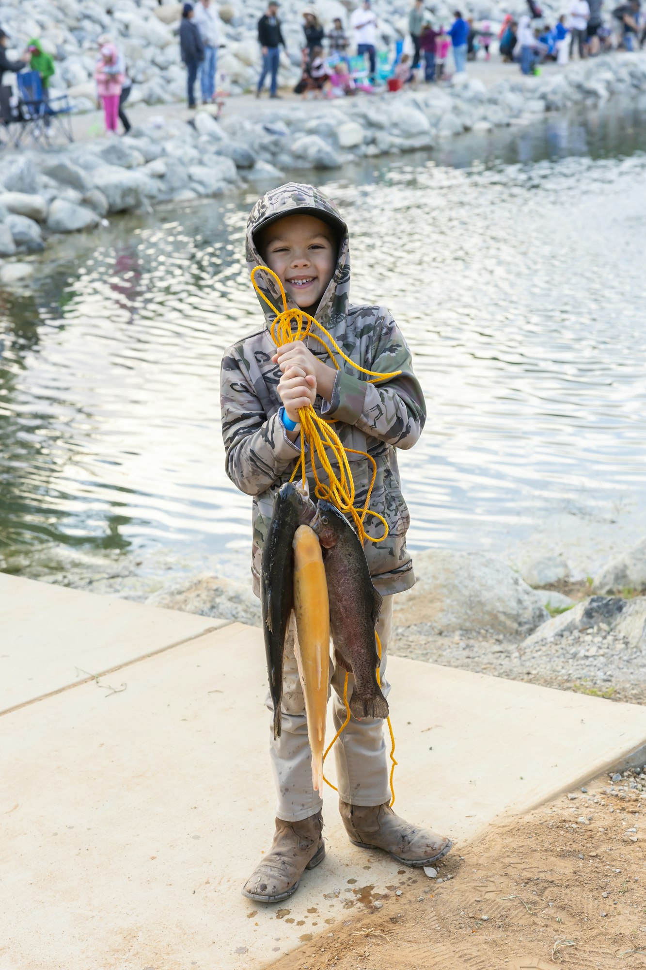 A child in camouflage gear holds a rope with several fish by a rocky lakeside.