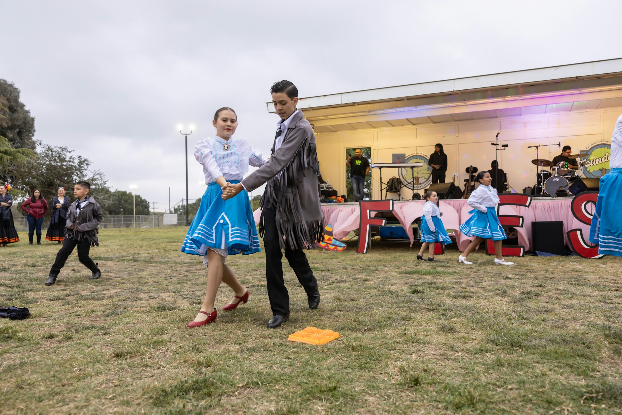 Dancers in traditional costumes perform outdoors, with a band and audience in the background.