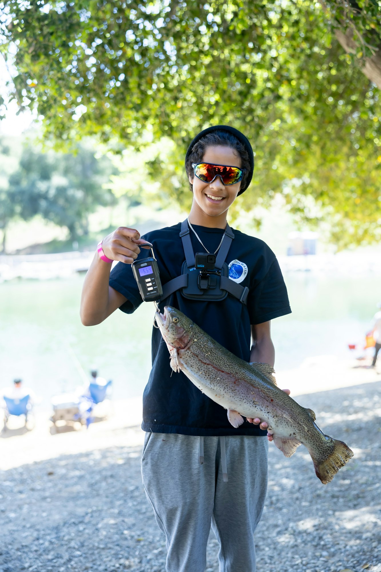 Person holding a large fish with a digital scale, wearing sunglasses and headphones, outdoors near a body of water.